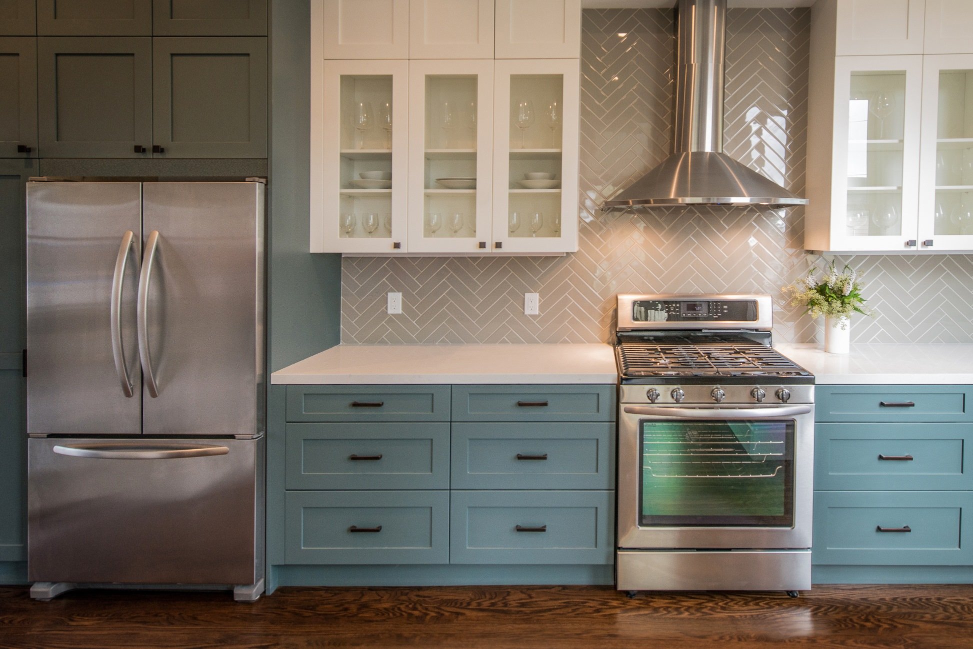 Kitchen with stainless steel refrigerator, white cabinets with glass doors, gray herringbone tile backsplash, stainless steel stove and vent hood, and a white countertop with a vase of flowers.