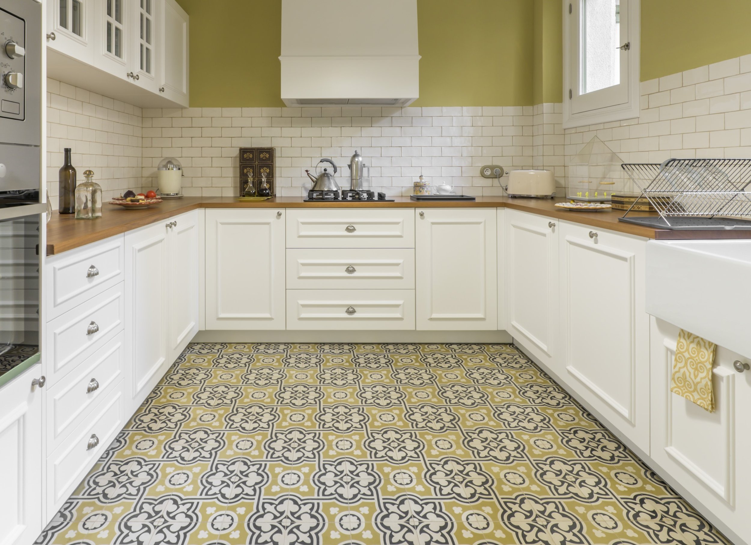 A kitchen with white cabinets, a wooden countertop, and a black and white patterned tile floor. There are bottles, jars, and kitchen appliances on the counter, along with a dish rack and a window.