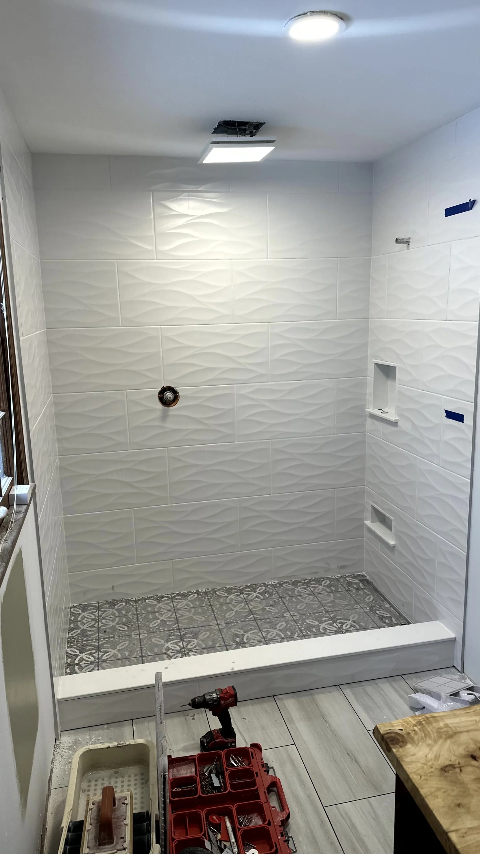 Bathroom shower with new white textured tiles, a recessed shelf, an opening for a showerhead, and a ceiling light fixture, still under renovation.
