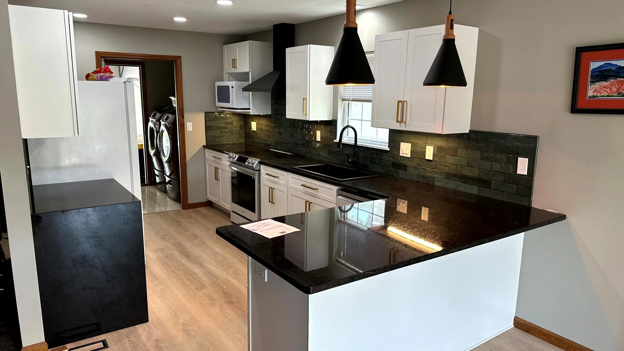 Modern kitchen with black countertops, white cabinets with gold handles, black backsplash, black pendant lights, and a window above the sink.