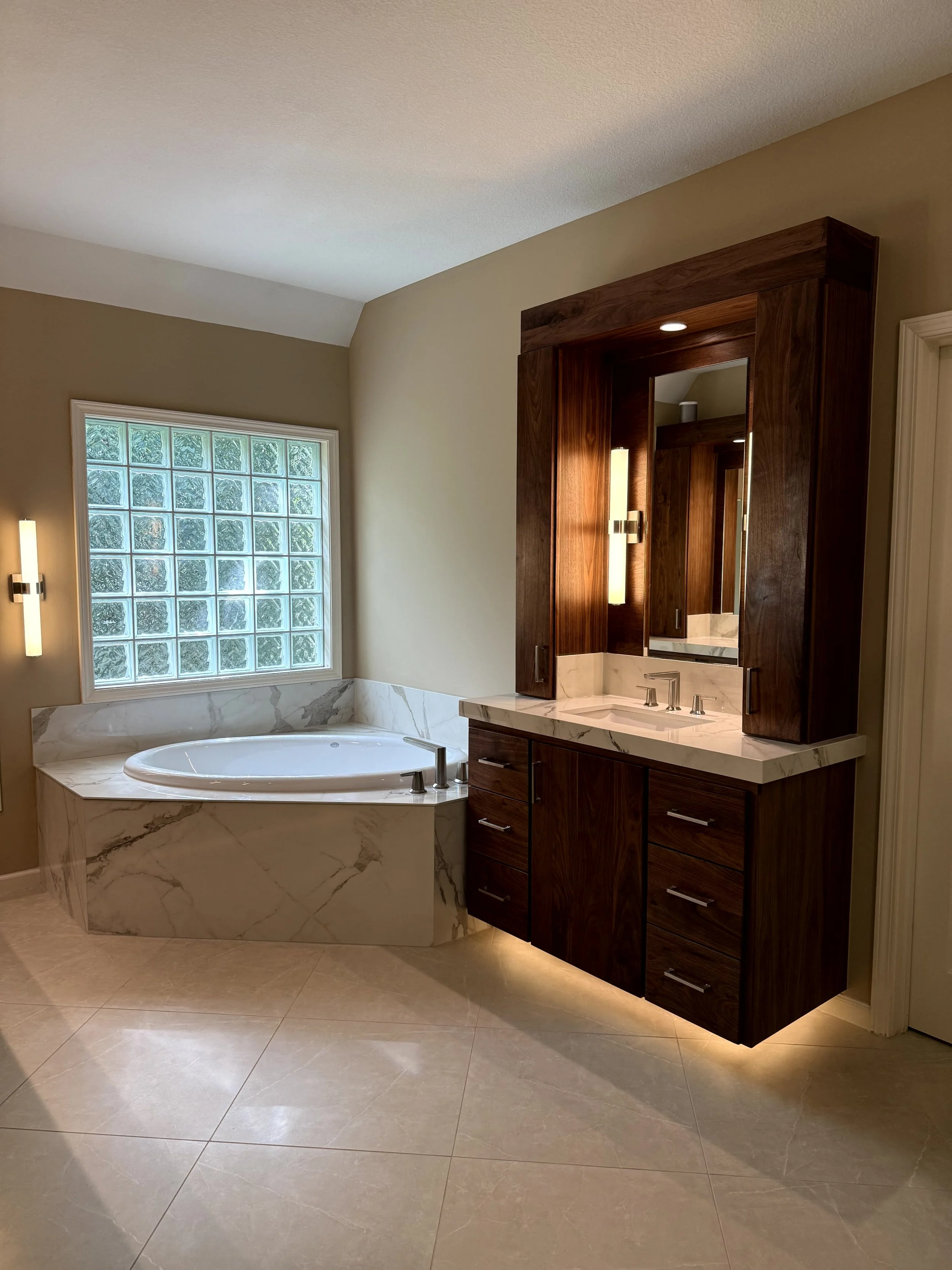 A bathroom with a marble tub, a glass block window, and a dark wood vanity with a mirror and sink.