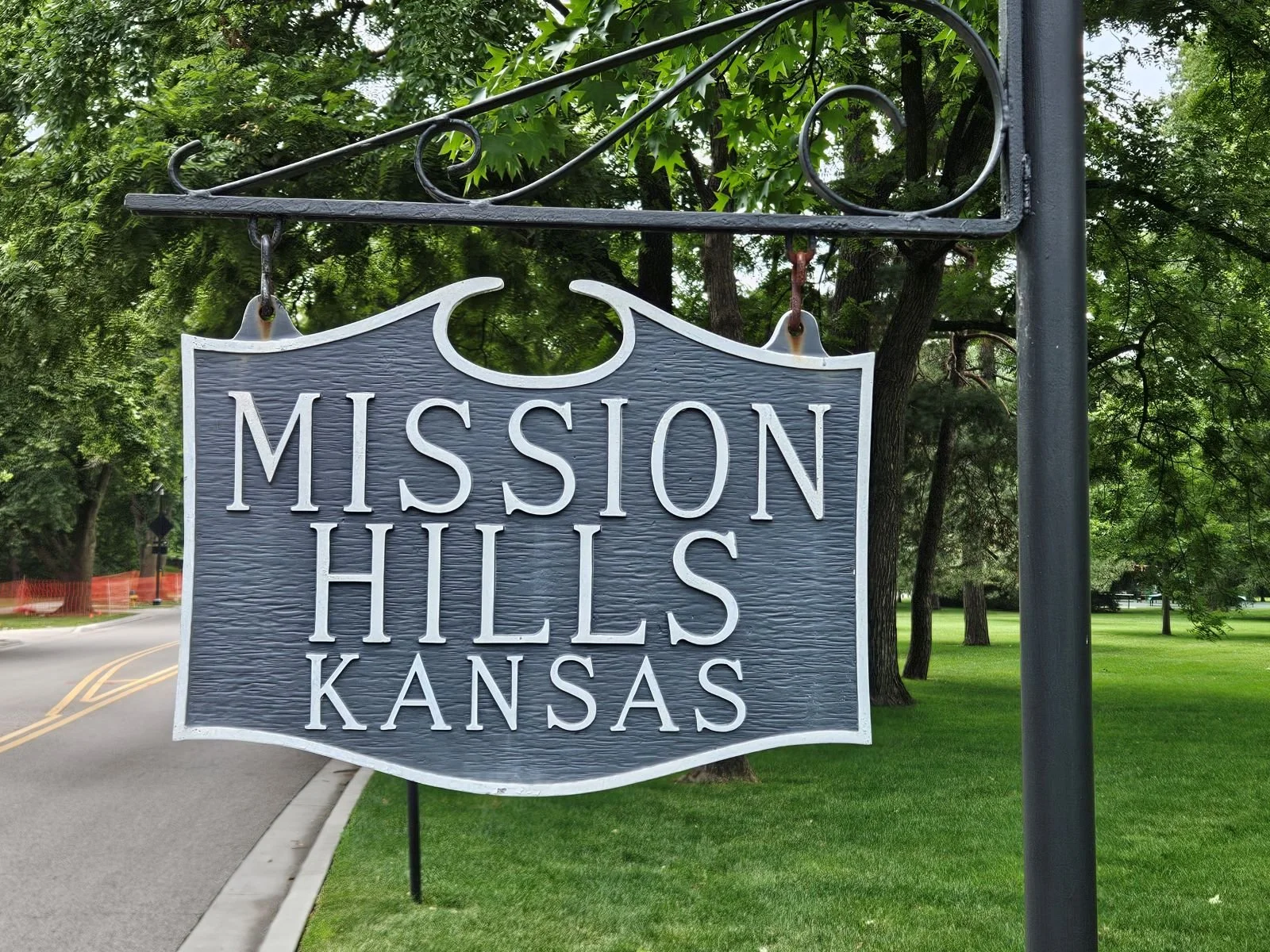 A hanging sign that says "Mission Hills Kansas" in white letters on a dark textured background, with a decorative black iron frame above it, set in a park with trees and a lawn in the background.