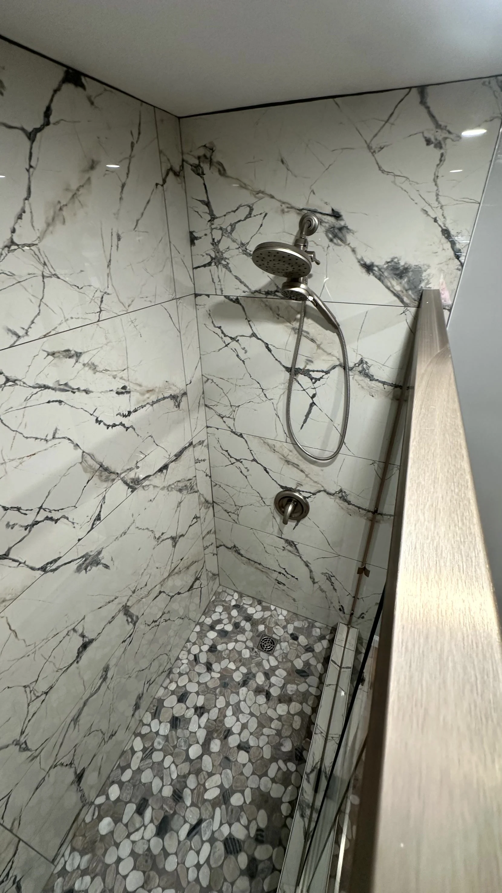 Shower with marble walls, a handheld showerhead, a wooden privacy partition, and pebble-style floor tiles.
