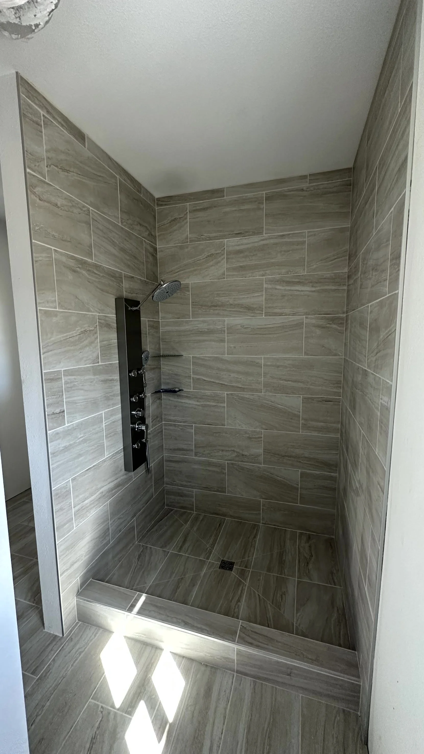 A modern walk-in shower with beige marble tiles, a black shower panel with multiple jets, a handheld showerhead, and a drain on the floor.