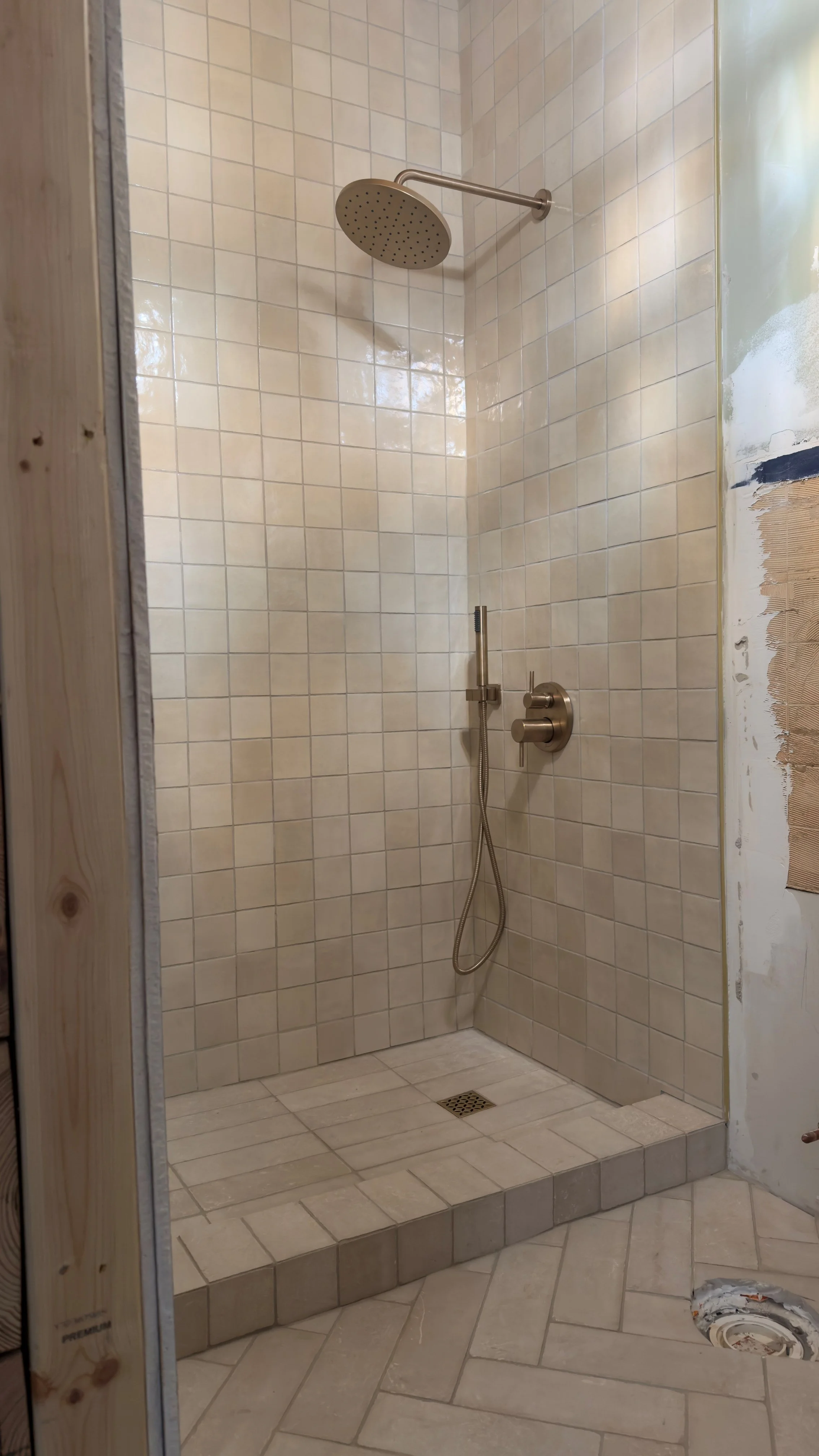 Construction site shower with beige tiles, a round rainfall showerhead, and a handheld shower attached to the wall, with unfinished wall and exposed drywall nearby.