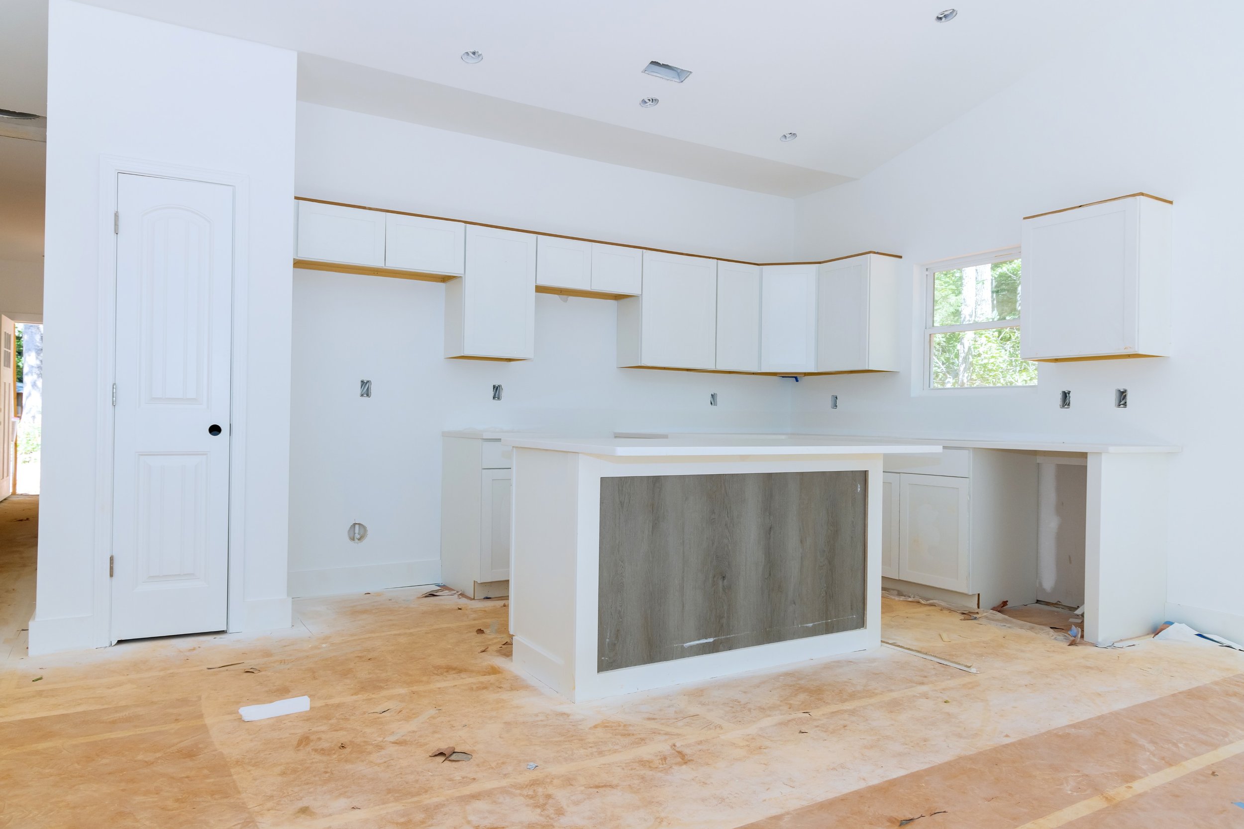 Kitchen under construction with white cabinets and an island, unfinished flooring, and a window showing trees outside.