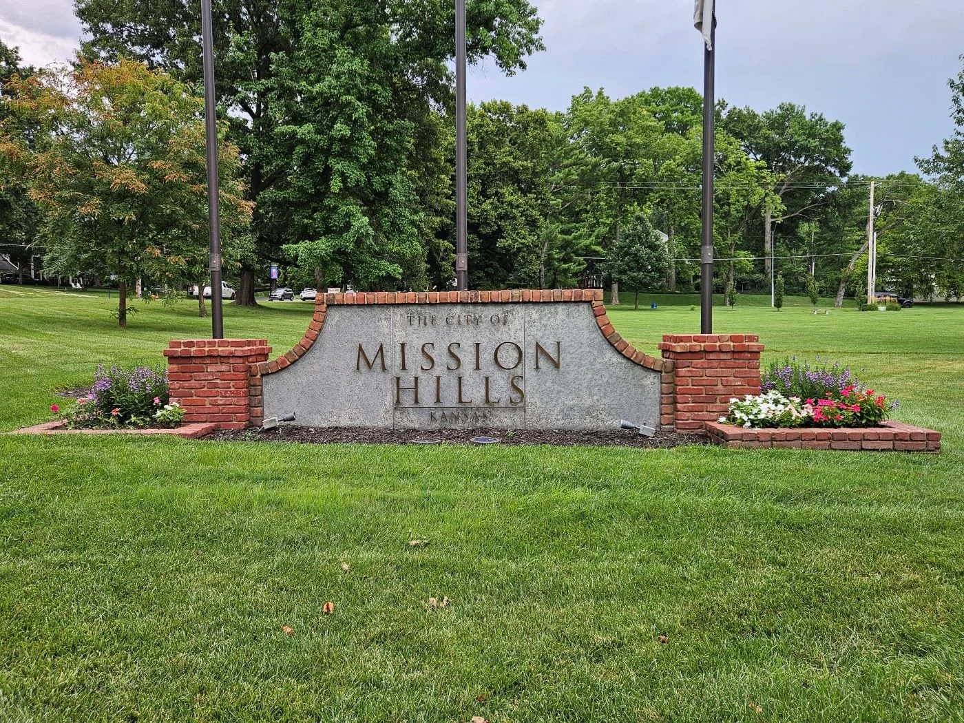 Sign reading "The City of Mission Hills Kansas" on a brick and stone monument, surrounded by grass and flower beds with trees in the background.