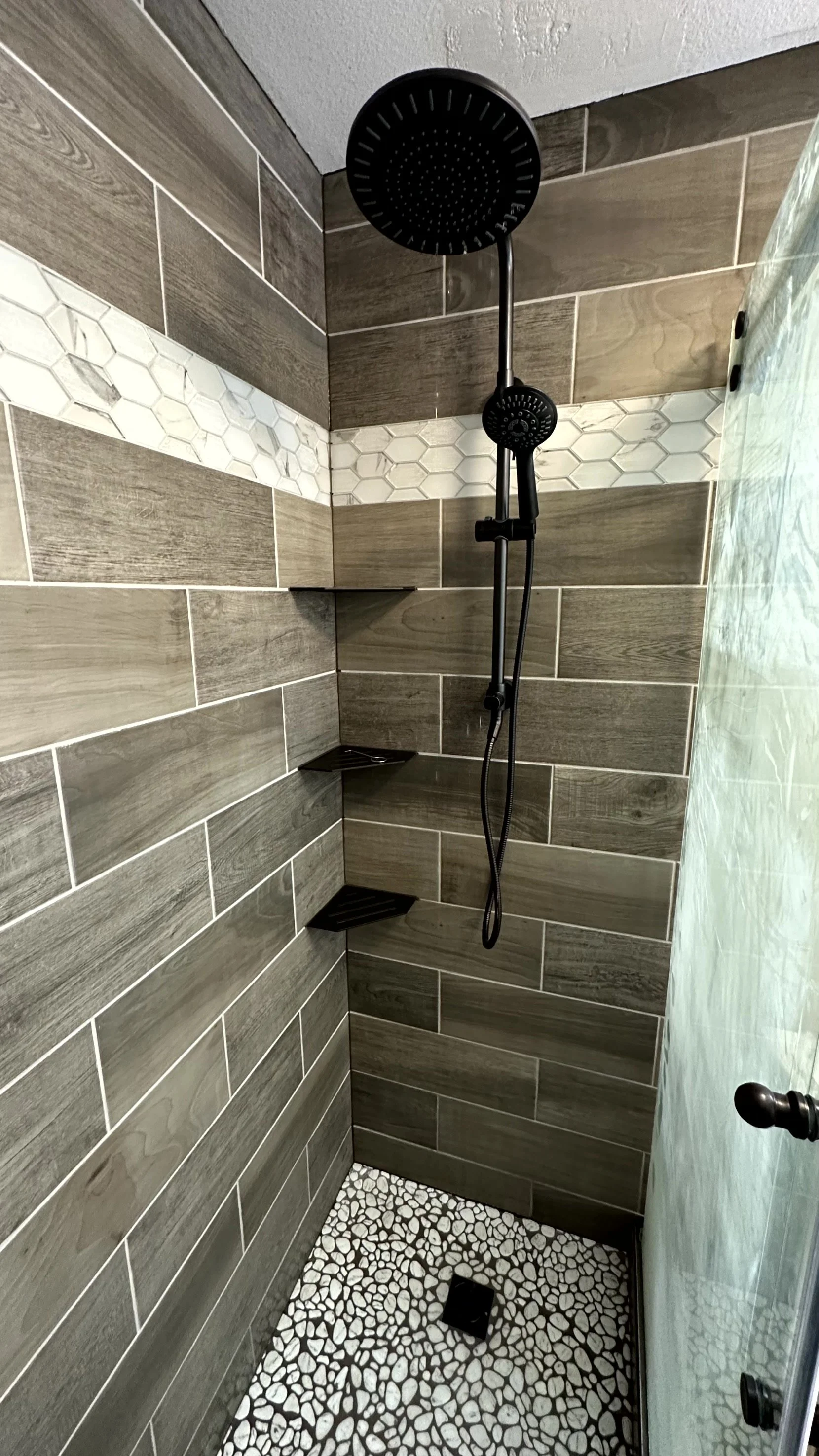 Shower with black round shower head, handheld shower wand, wooden wall tiles, pebble mosaic floor, small triangular corner shelves, partially glass shower door, and a black door handle.