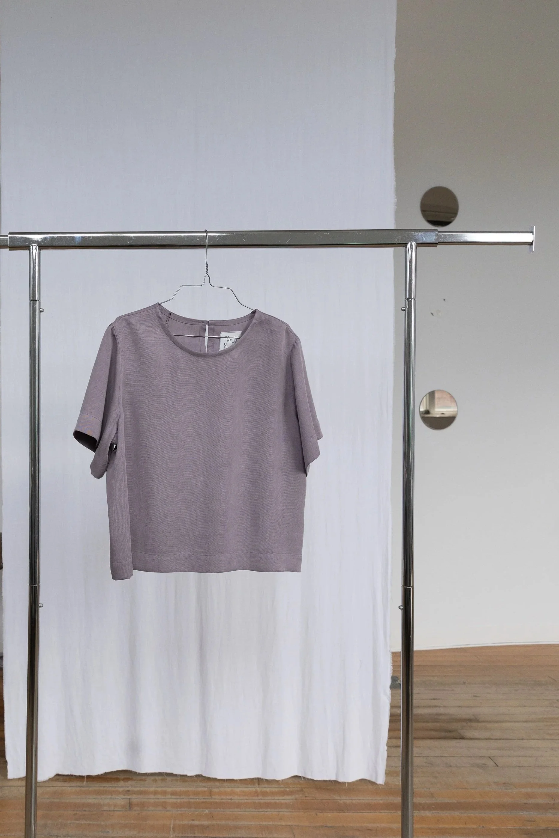 Une robe gris clair suspendue à un porte-vêtements en métal, avec un fond de rideau blanc et un mur gris avec des trous circulaires.