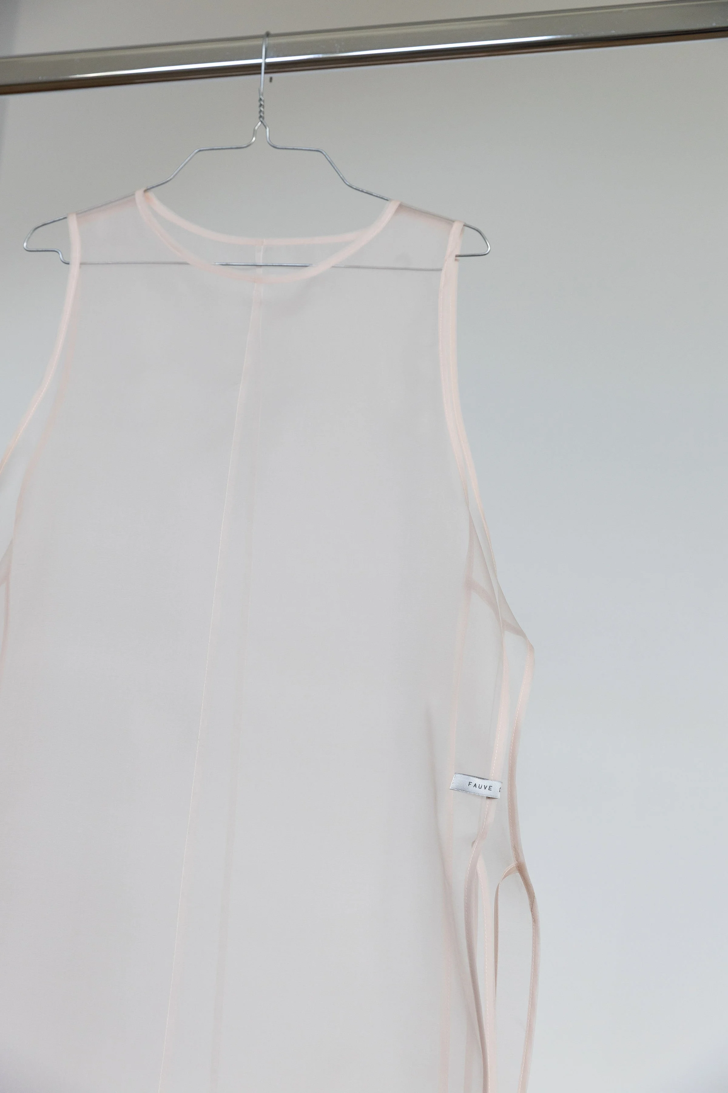 Robe en tissu léger de couleur blanche ou très claire suspendue sur un cintre en métal. La robe a des bretelles fines et un petit étiquette avec la marque 'FAUVE' attachée à la couture en bas à droite.