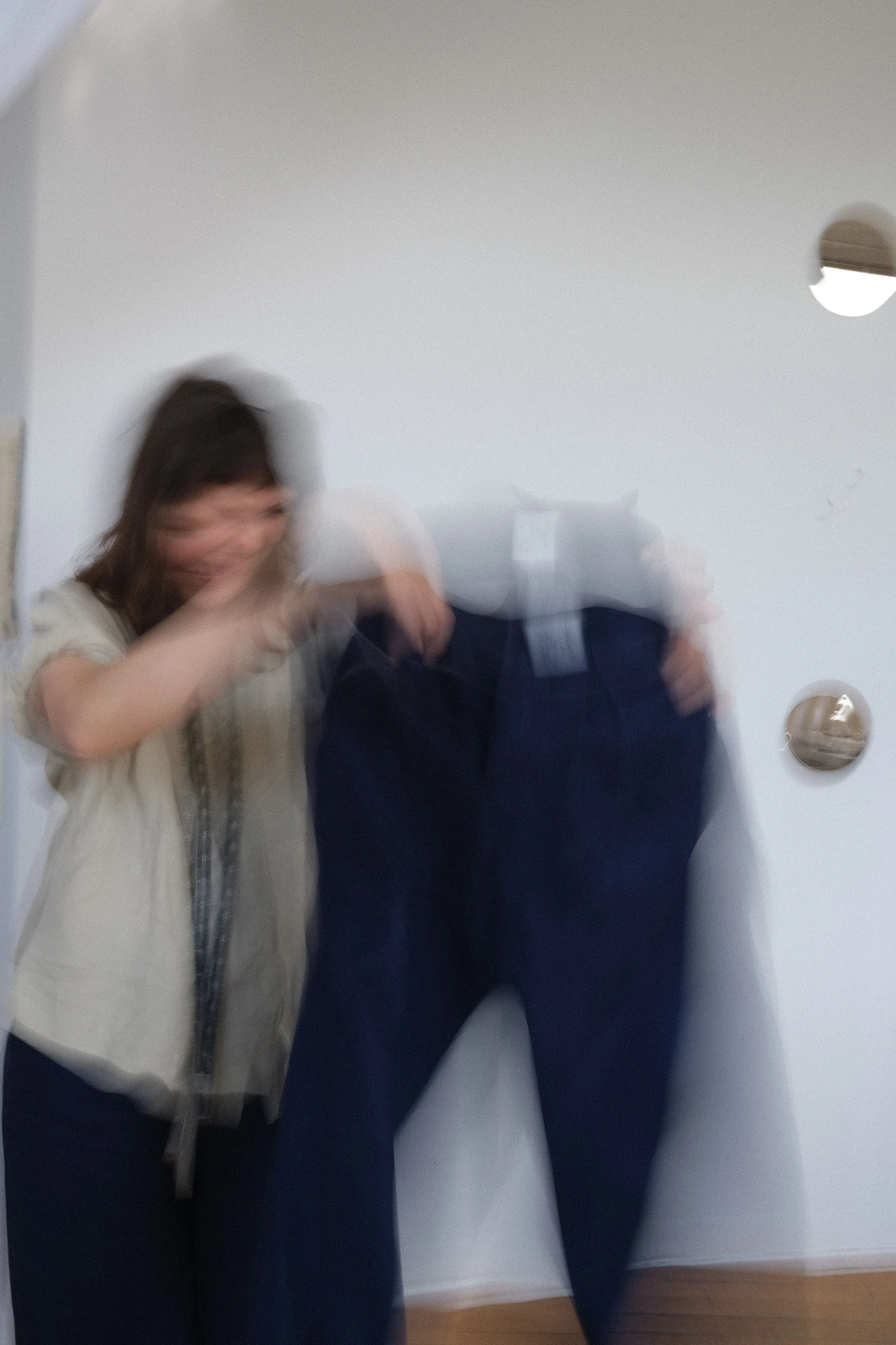 Reflet flou d'une personne souriante avec des cheveux bruns, portant une chemise beige avec une écharpe à motifs, dans un miroir situé dans une pièce avec murs blanc et sol en bois.