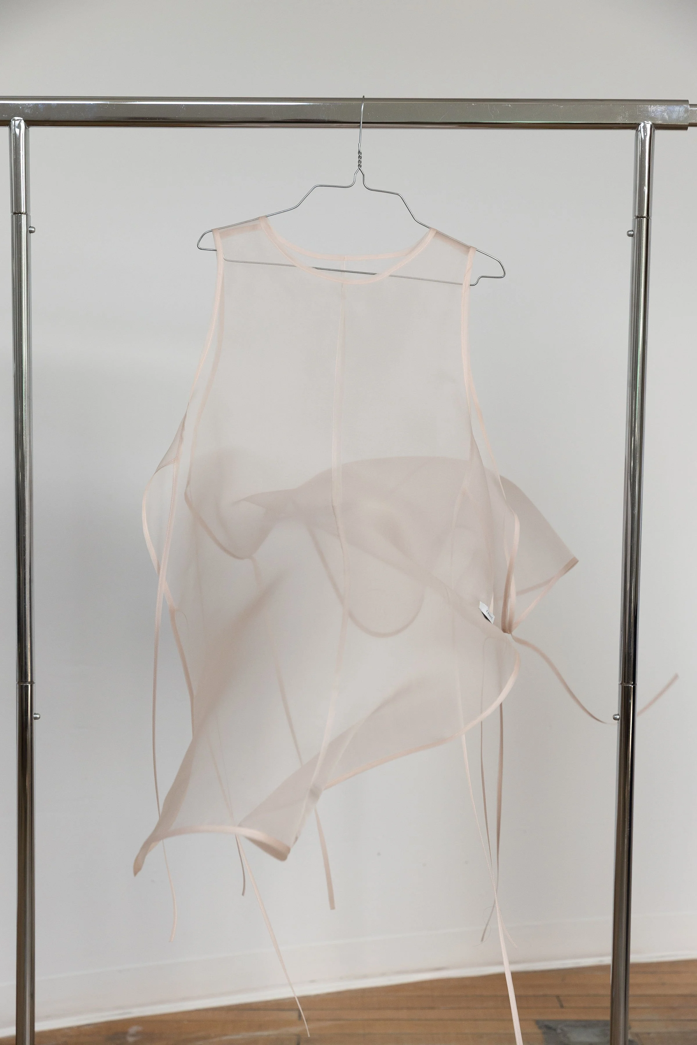 Une robe en tissu transparent suspendue à une chaise en métal, dans une pièce avec un plancher en bois et un mur blanc.