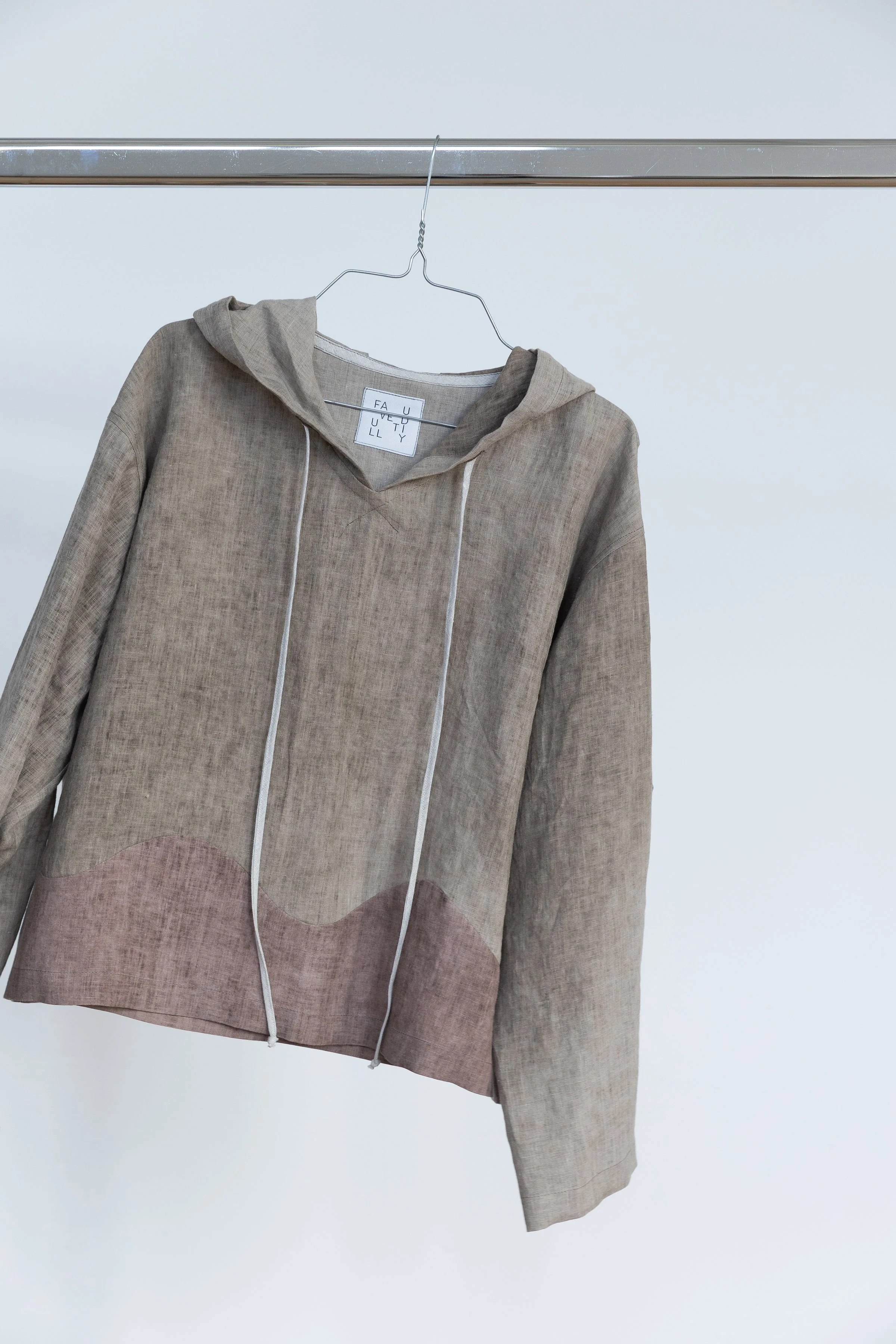 Veste en tissu beige avec capuche, suspendue sur une branche métallique, fond blanc.