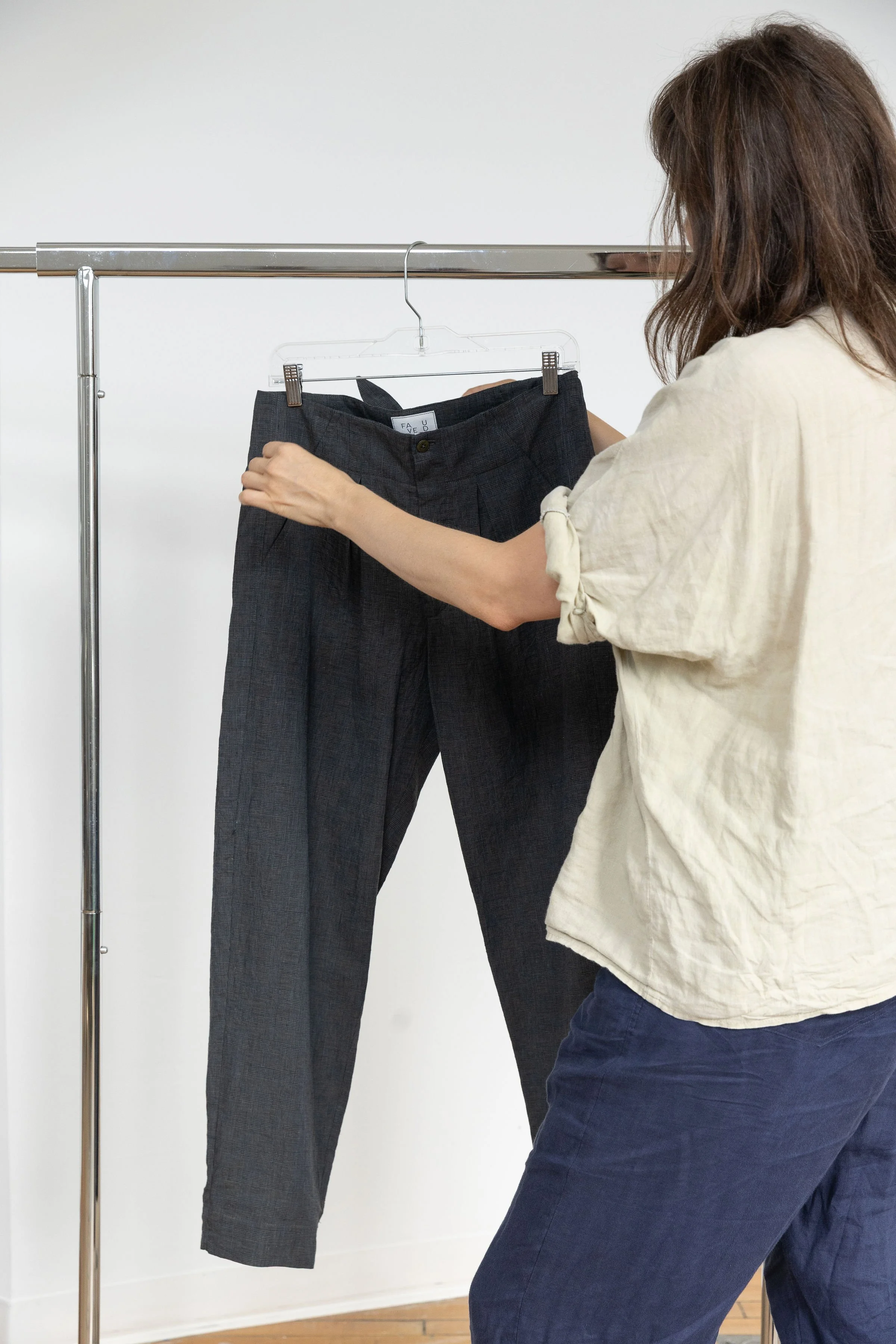Une femme qui essaie un pantalon noir à carreaux dans une boutique ou un studio, utilisant un portemanteau en verre suspendu pour accrocher le vêtement.