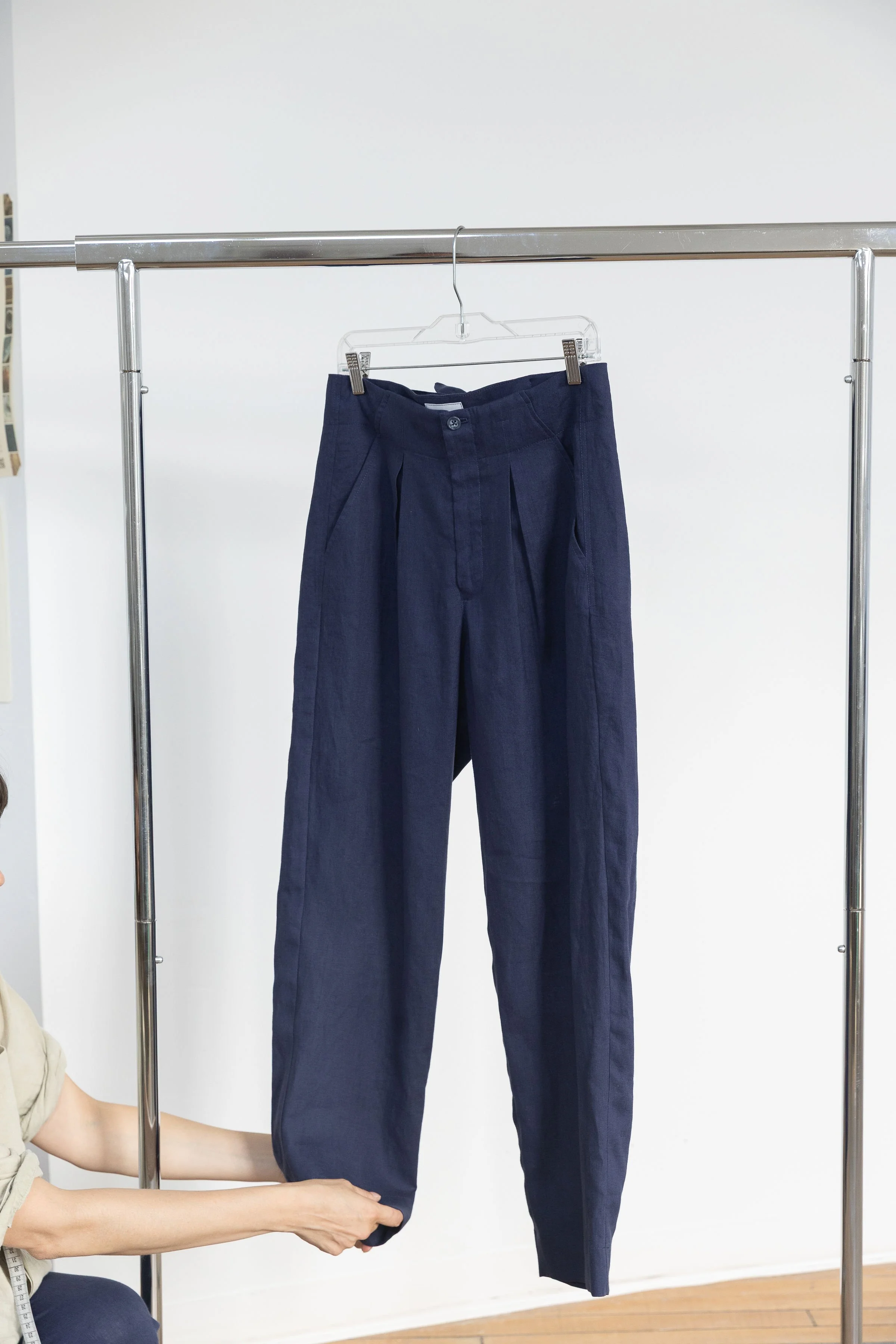 Un pantalon bleu foncé suspendu sur un portemanteau. Une personne tient le pantalon au niveau de la cheville avec ses mains dans un intérieur. La scène est bien éclairée avec un mur blanc en arrière-plan.