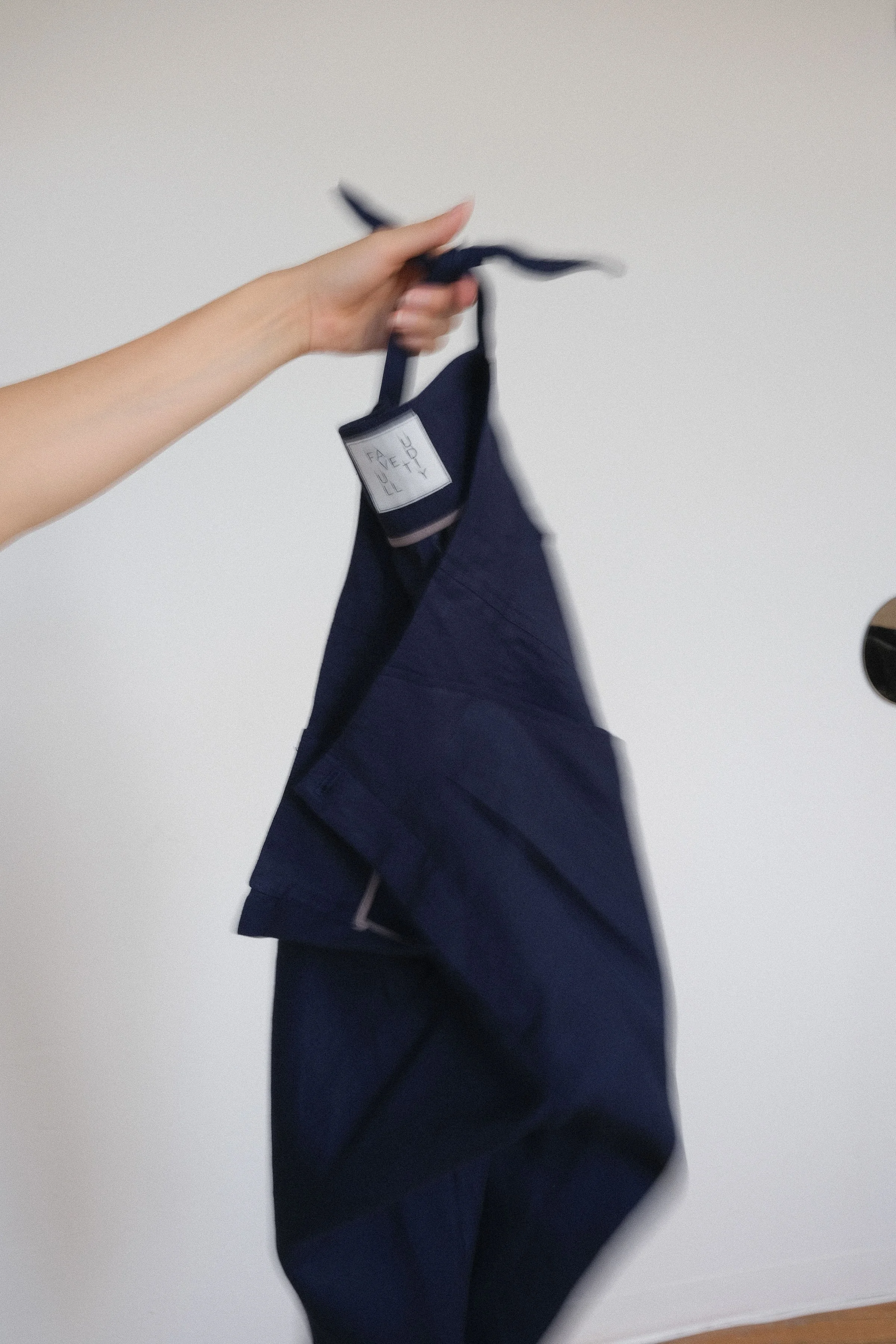Une main tient un vêtement bleu, probablement un pantalon ou un short, contre un mur blanc.