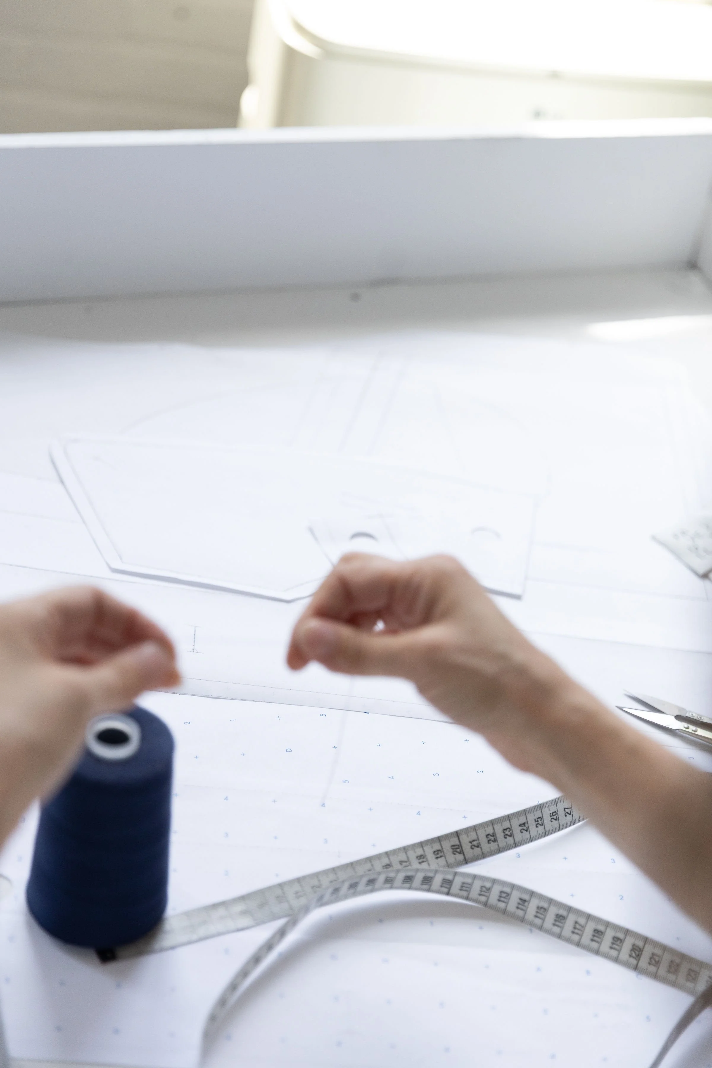 Personne en train de manipuler un patron en papier pour la couture, avec des ciseaux, une règle de couture et un tableau de tracés sur la table de travail.
