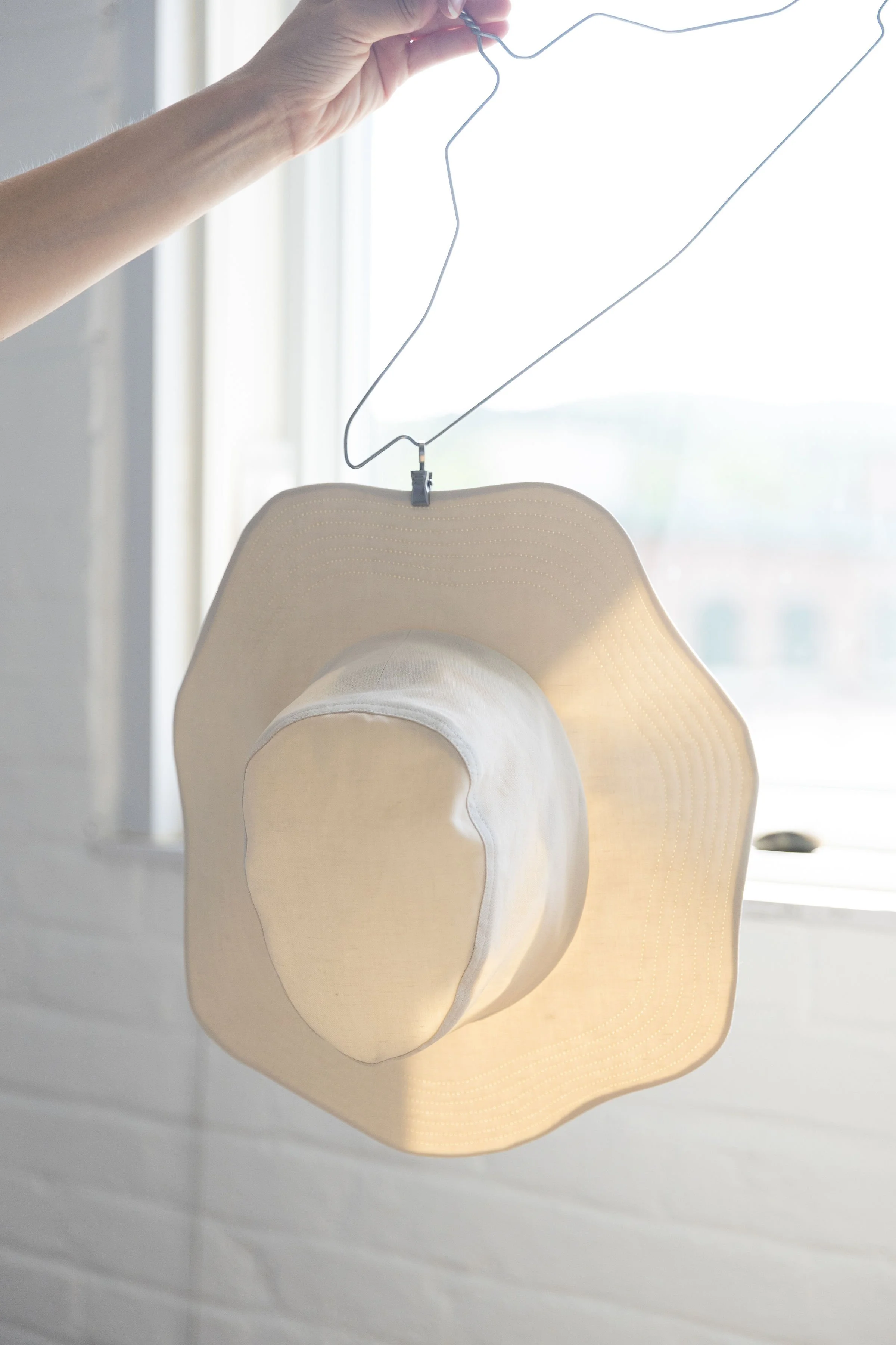 Un chapeau beige suspendu à un cintre en fil métallique, situé près d'une fenêtre avec une lumière naturelle qui illumine la scène.