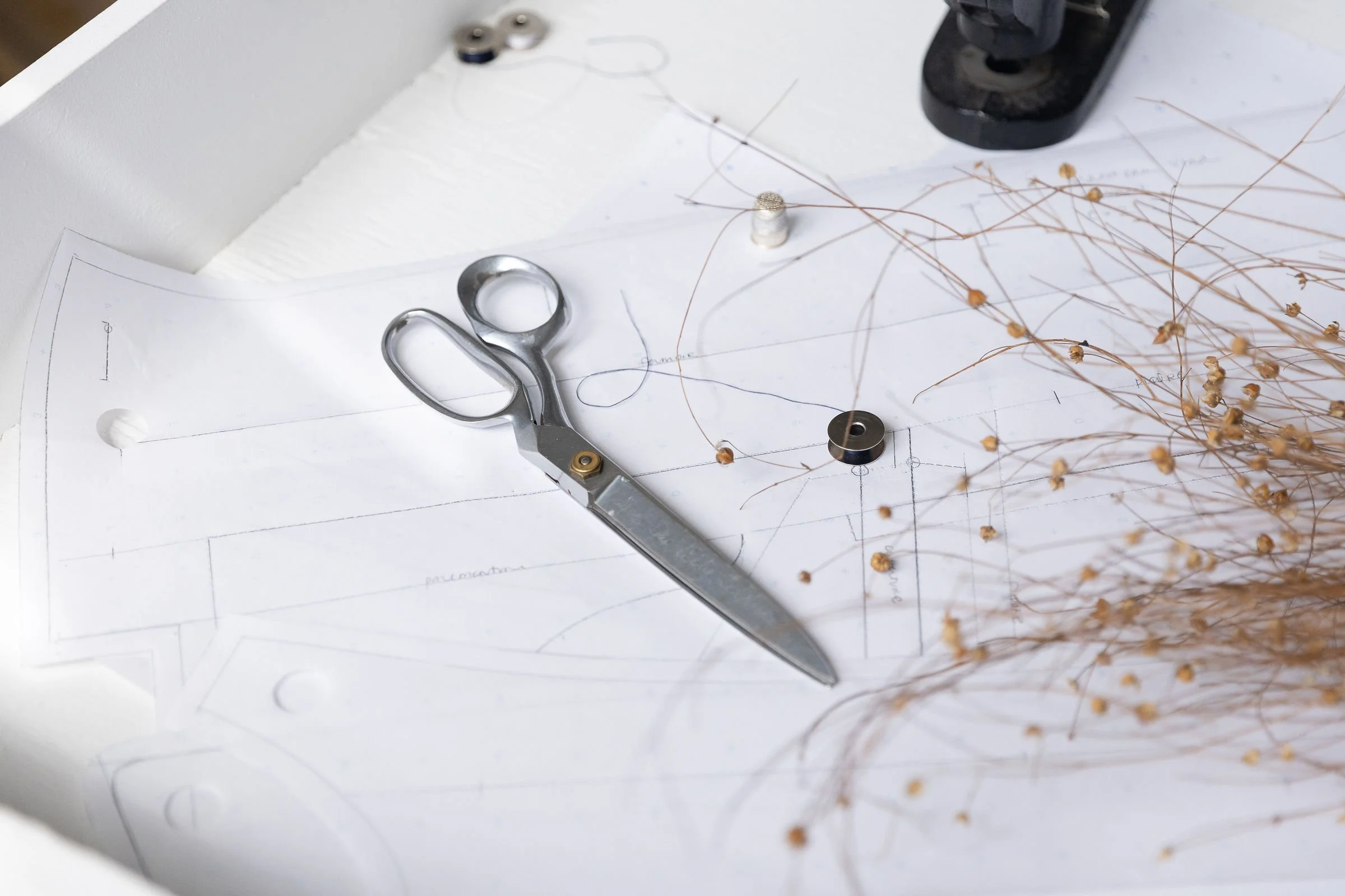 Une paire de ciseaux, des échantillons de fil d'or, un tournevis, et un dessin technique sur une feuille de papier avec des tracés et des annotations.