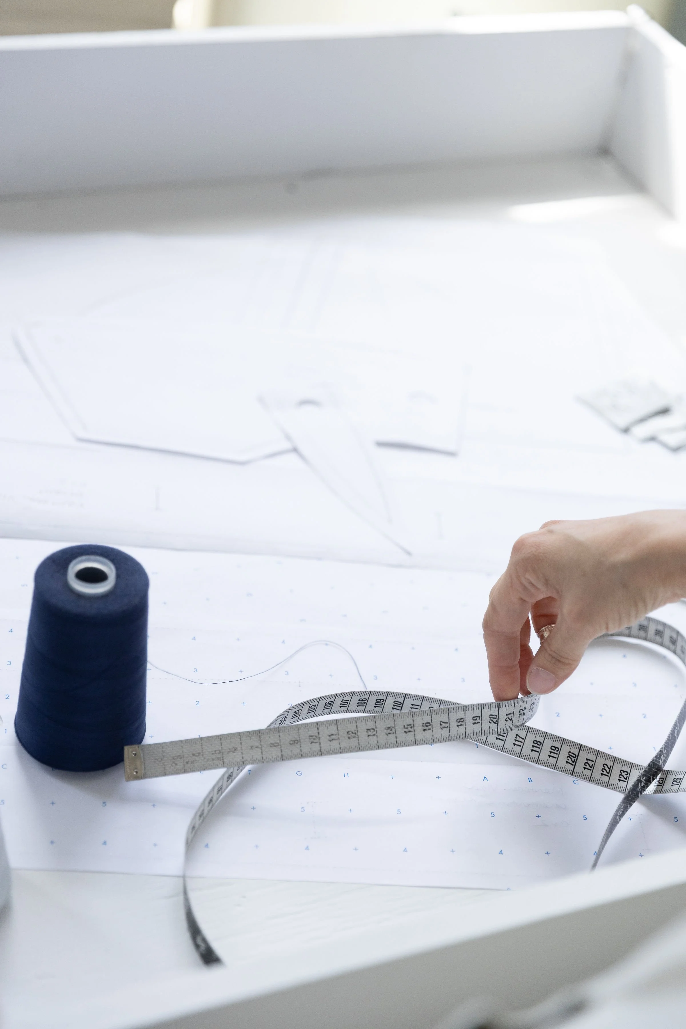 Un bobine de fil bleu, un ruban à mesurer déroulé, un plan de couture et une main ajustant le ruban sur un bureau