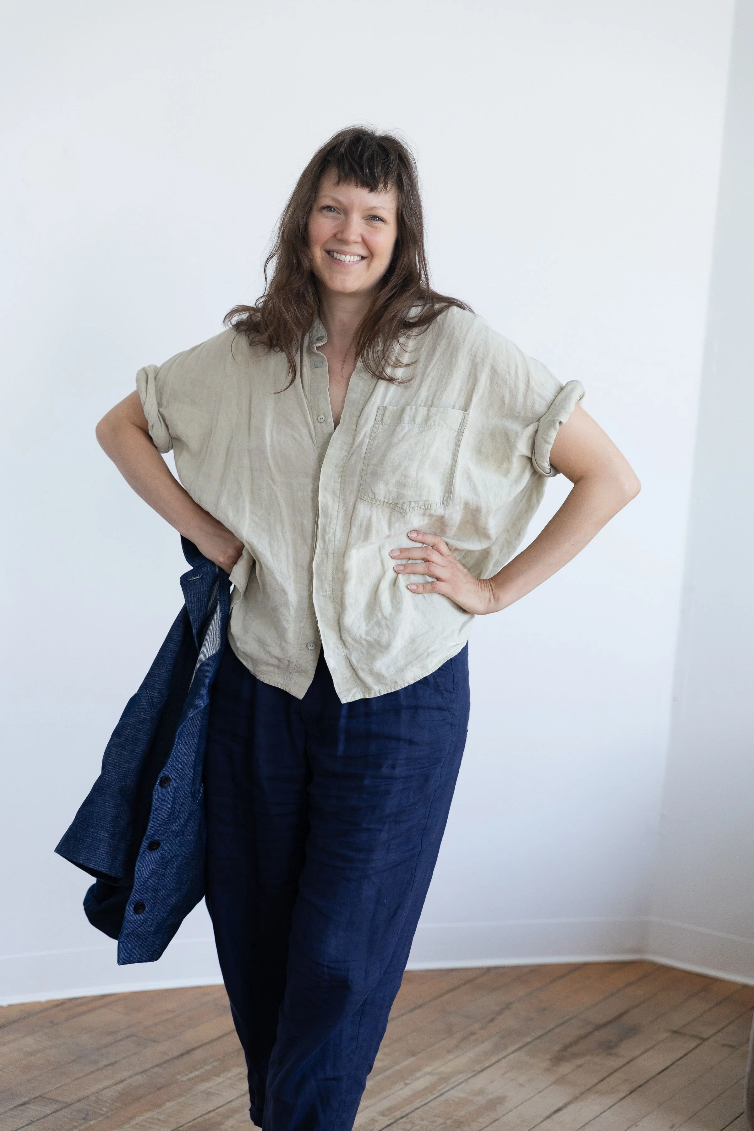 Une femme souriante avec des cheveux foncés et bouclés, portant une chemise beige à manches retroussées, et tenant un manteau en jean sur son bras gauche, debout dans une pièce avec un mur blanc et un plancher en bois.