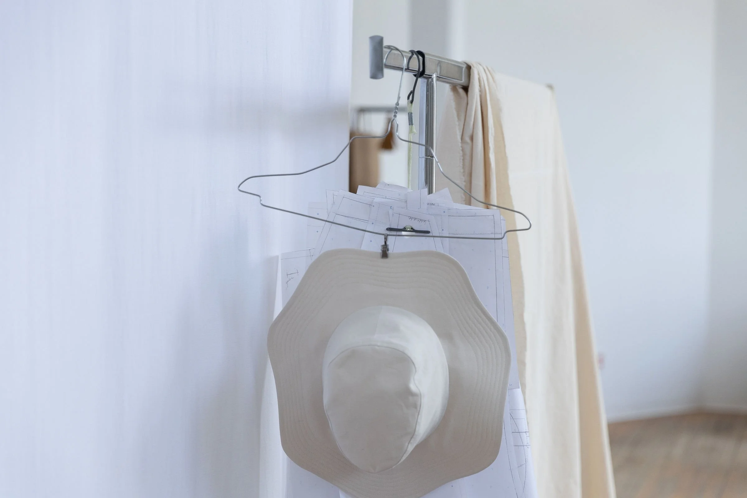 Panier de vêtements suspendus avec des papiers, un chapeau blanc et un vêtement beige ou crème suspendus dans une pièce avec mur blanc.