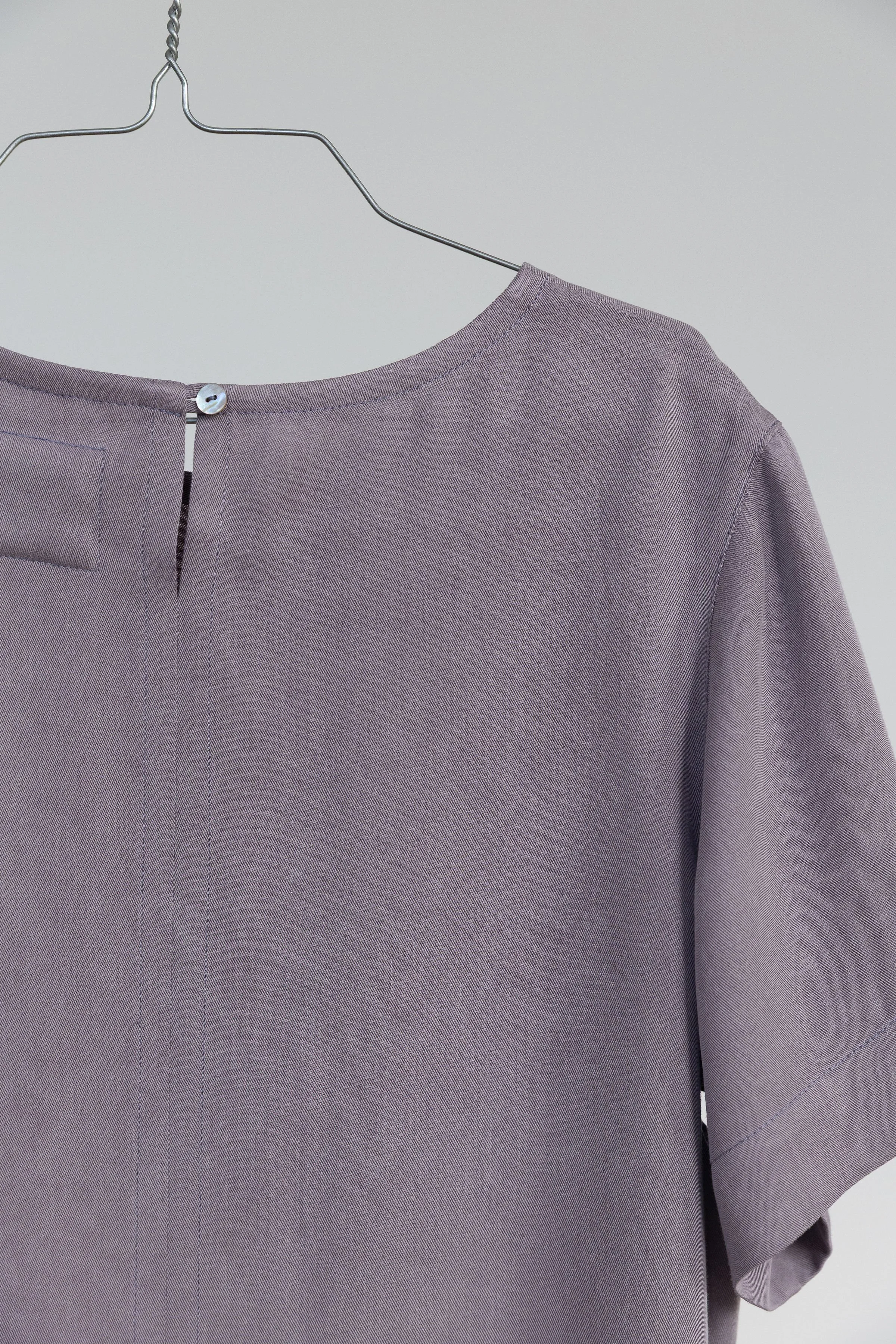 Veste violette avec un bouton blanc et un col rond suspendue sur un cintre en métal dans un fond neutre.