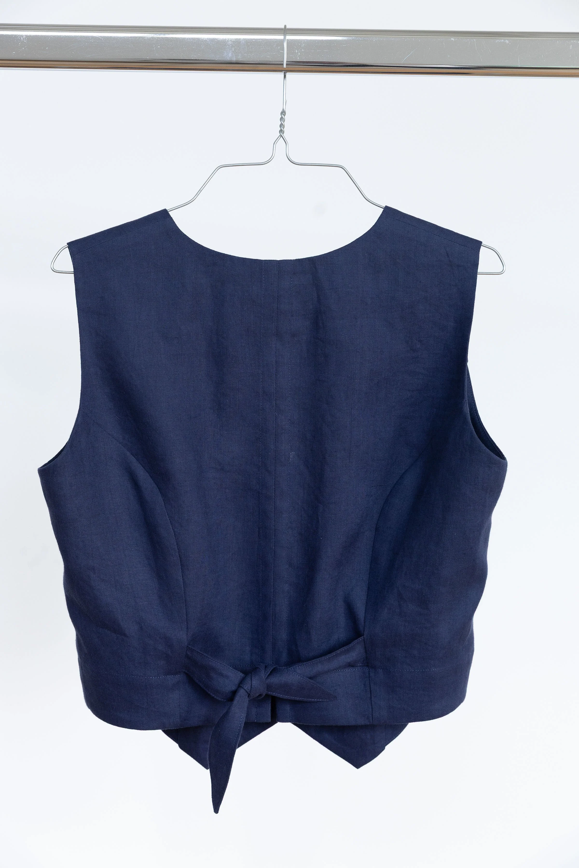 Veste bleue foncée avec un ceinture à nouer, suspendue sur un cintre en métal, contre un fond blanc.