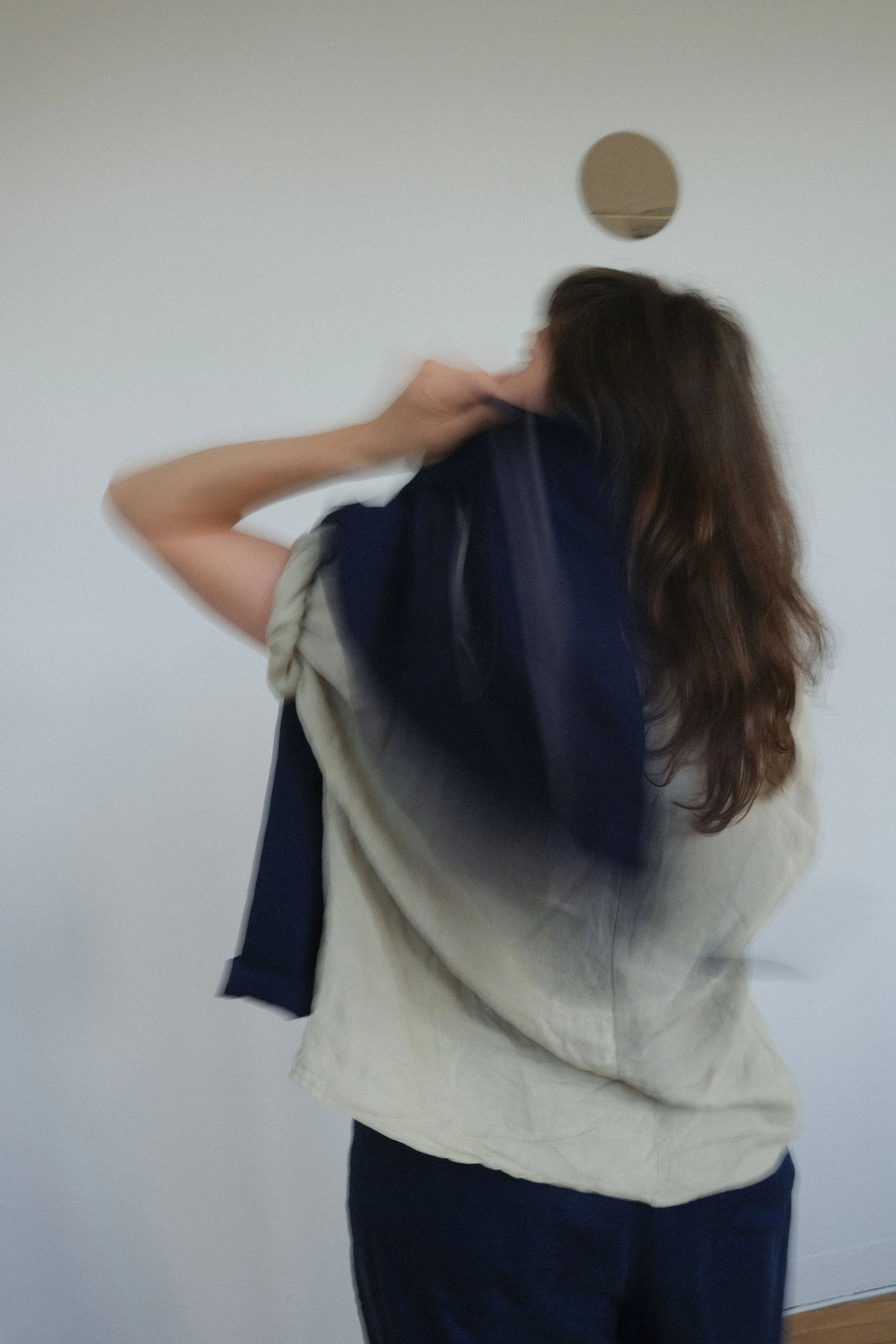 Une femme vue de dos, portant un sweat blanc, une veste bleue et un pantalon noir, face à un mur blanc.