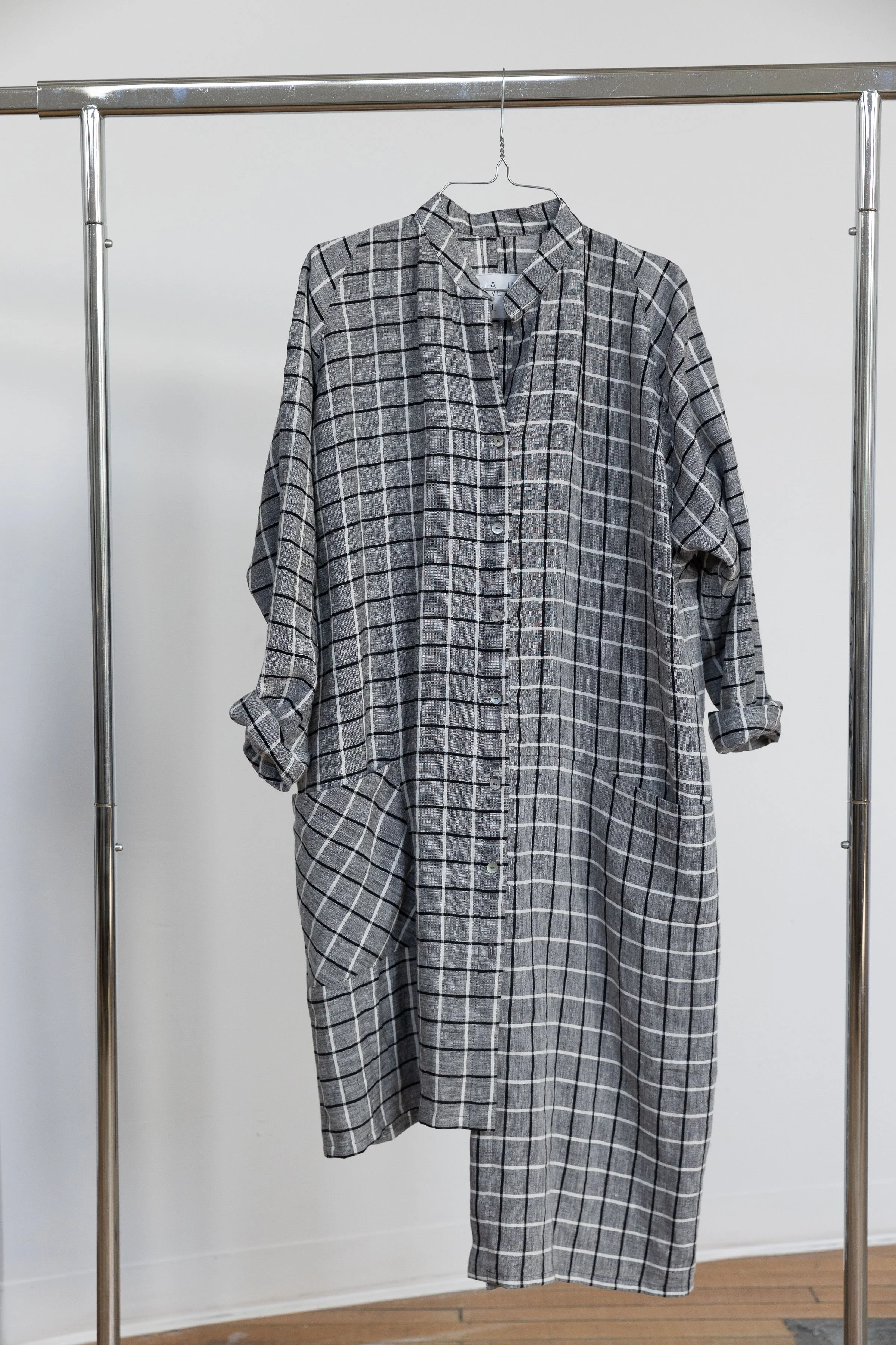 Chemise grise à carreaux noirs suspendue sur un portemanteau en métal, dans une pièce avec mur blanc et sol en bois.