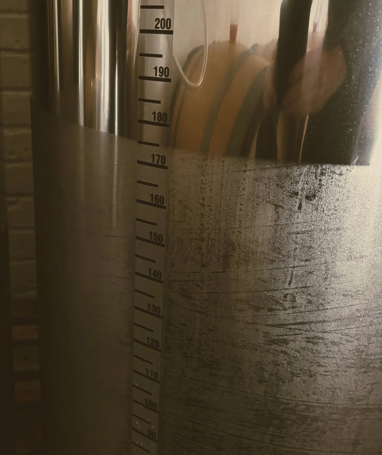 Close-up of a metallic measuring container or tank with measurement markings from 110 to 200 units, with a transparent tube or pipe attached, and a reflection of objects on its surface.