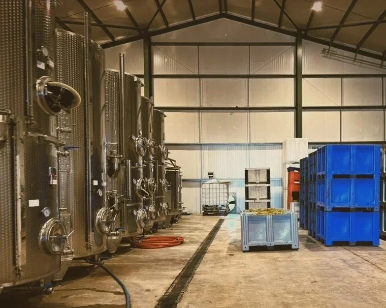 Industrial winery or brewery storage room with large stainless steel tanks on the left and blue plastic crates on the right, and a concrete floor.