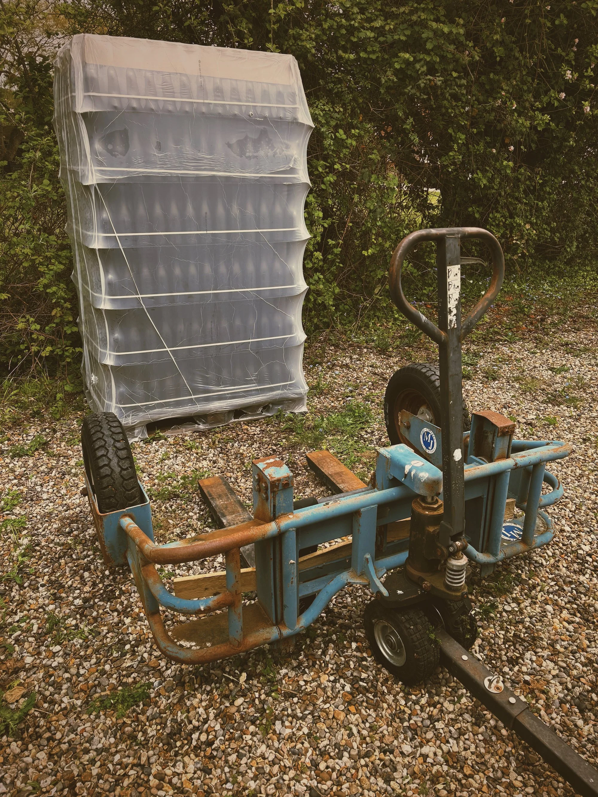 A rusted small vehicle or go-kart with missing parts, sitting on a gravel surface next to a damaged shelving unit covered with a plastic sheet, with green bushes and trees in the background.