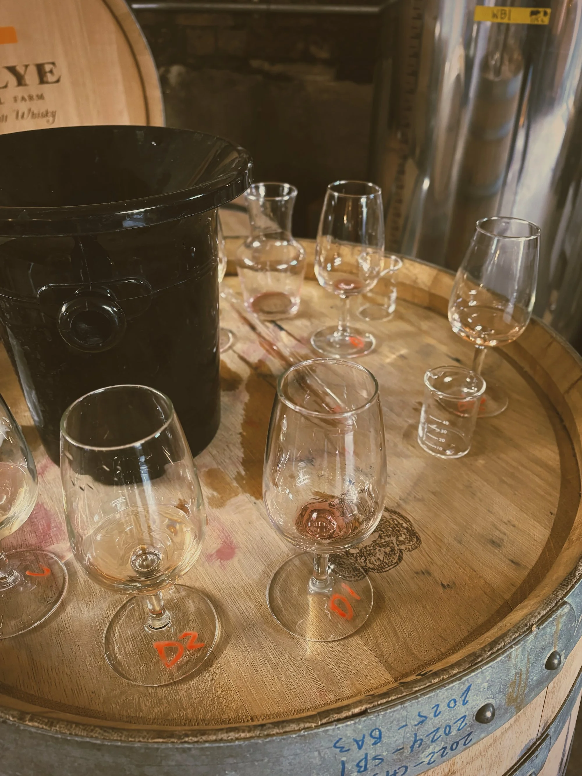 A wine aging batch with several empty glasses arranged on a wooden surface, a black bucket, and a metal wine barrel with handwritten notes.