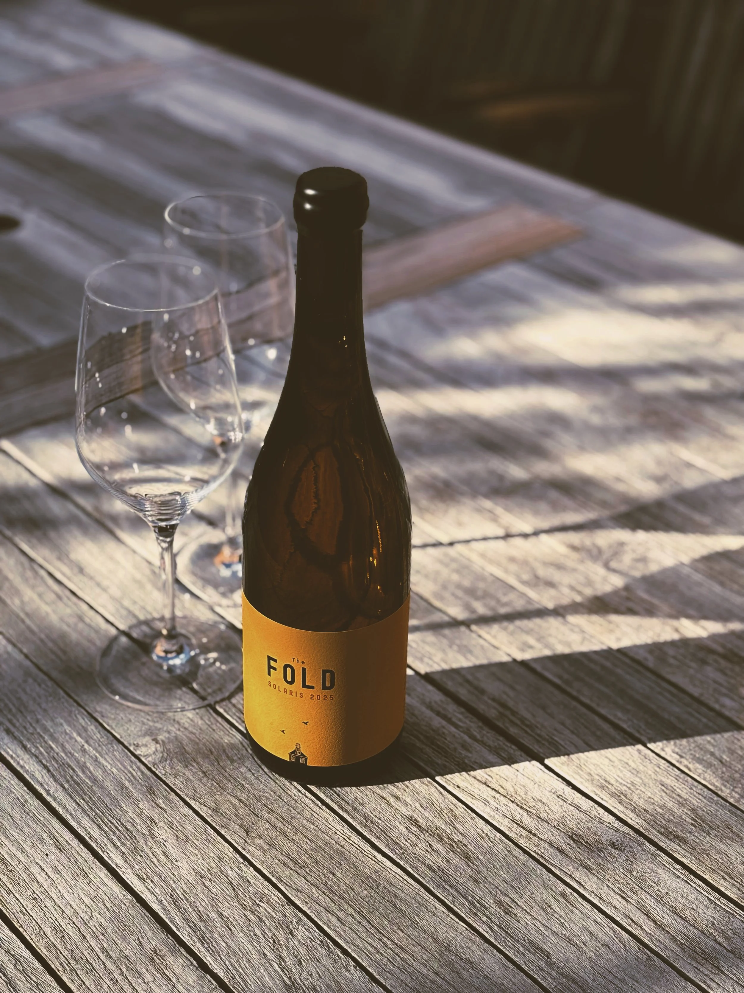 A brown beer bottle with a yellow label and two empty wine glasses on a weathered wooden surface outdoors.