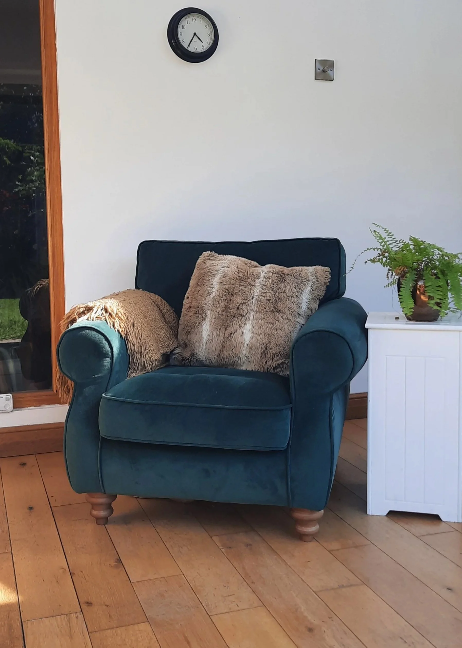 A teal upholstered armchair with wooden legs, placed on a wooden floor, with two faux fur pillows on it. To the right, there's a white side table with a potted fern. In the background, a white wall features a round black-framed clock showing 4:28 and a small metal hook. To the left, a wooden doorway looks out to an outside garden.