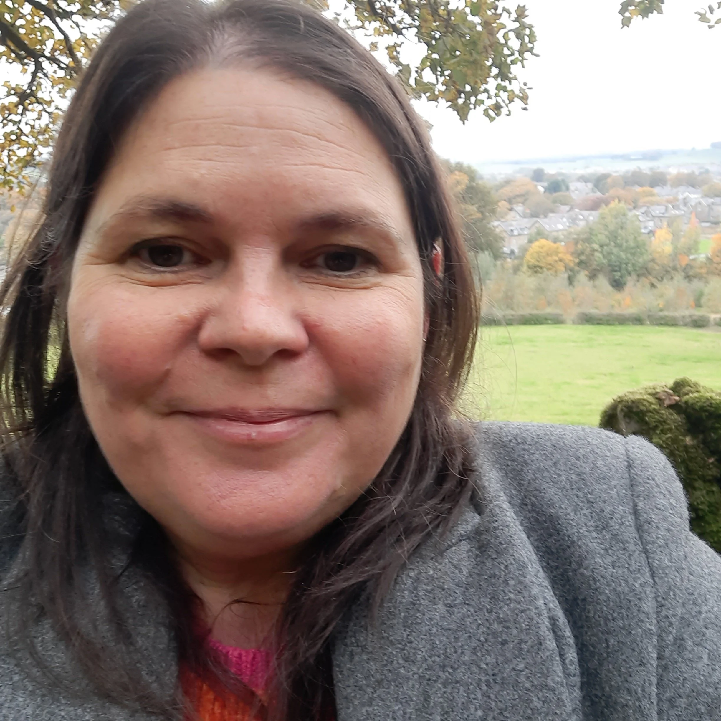 Close-up of a woman's face with brown hair, smiling outdoors near a tree and houses in the background.
