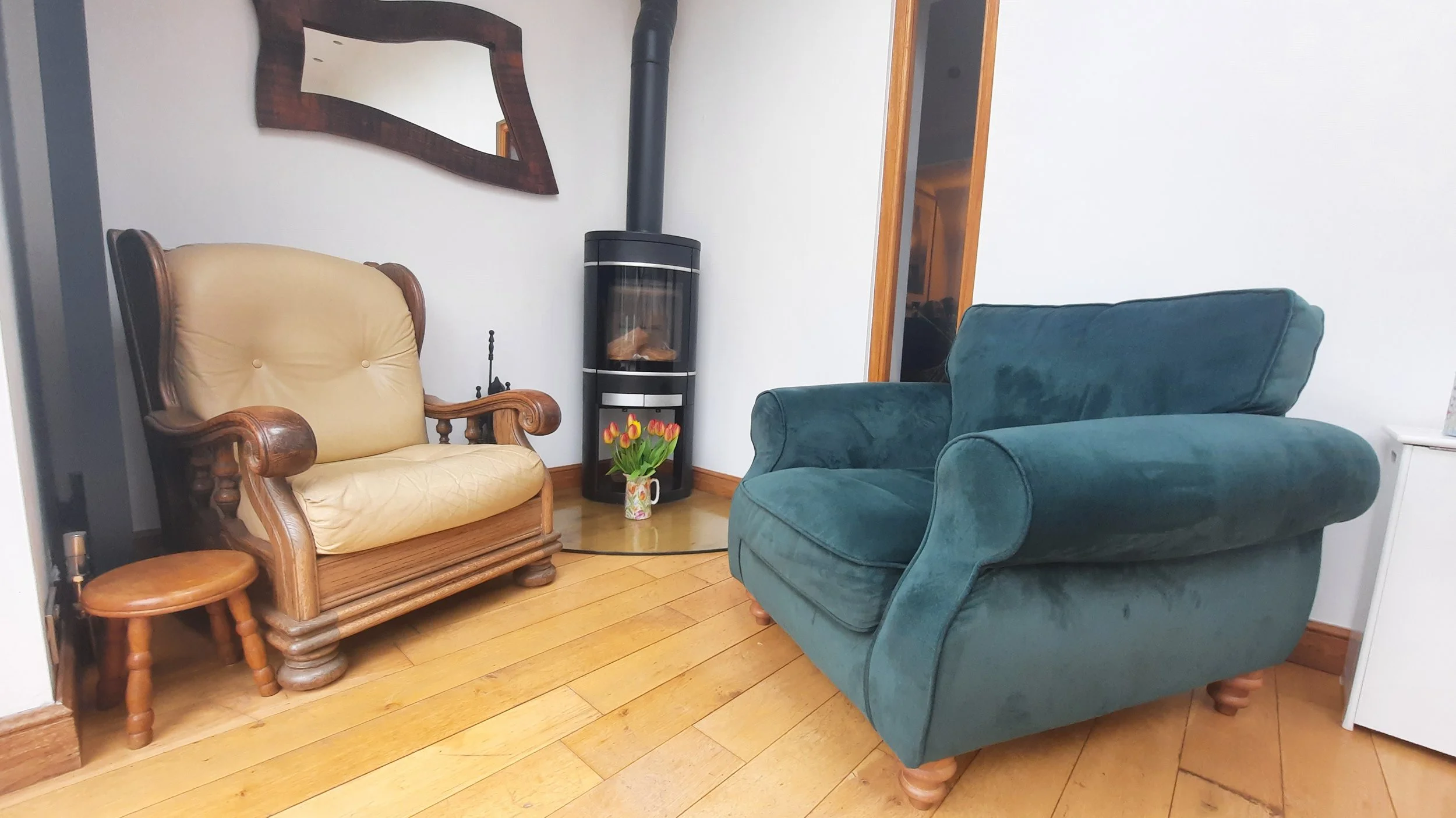 Living room with a beige armchair, a teal armchair, a small side table, a black pellet stove, and a vase of tulips on the floor.
