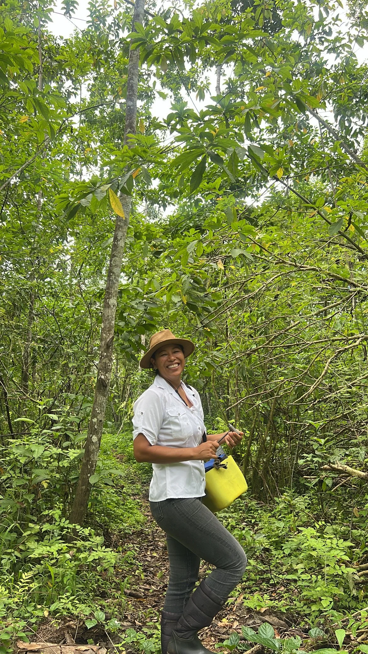 A woman smiling with a hat and white shirt in a dense green jungle, holding a small tool and a yellow container.