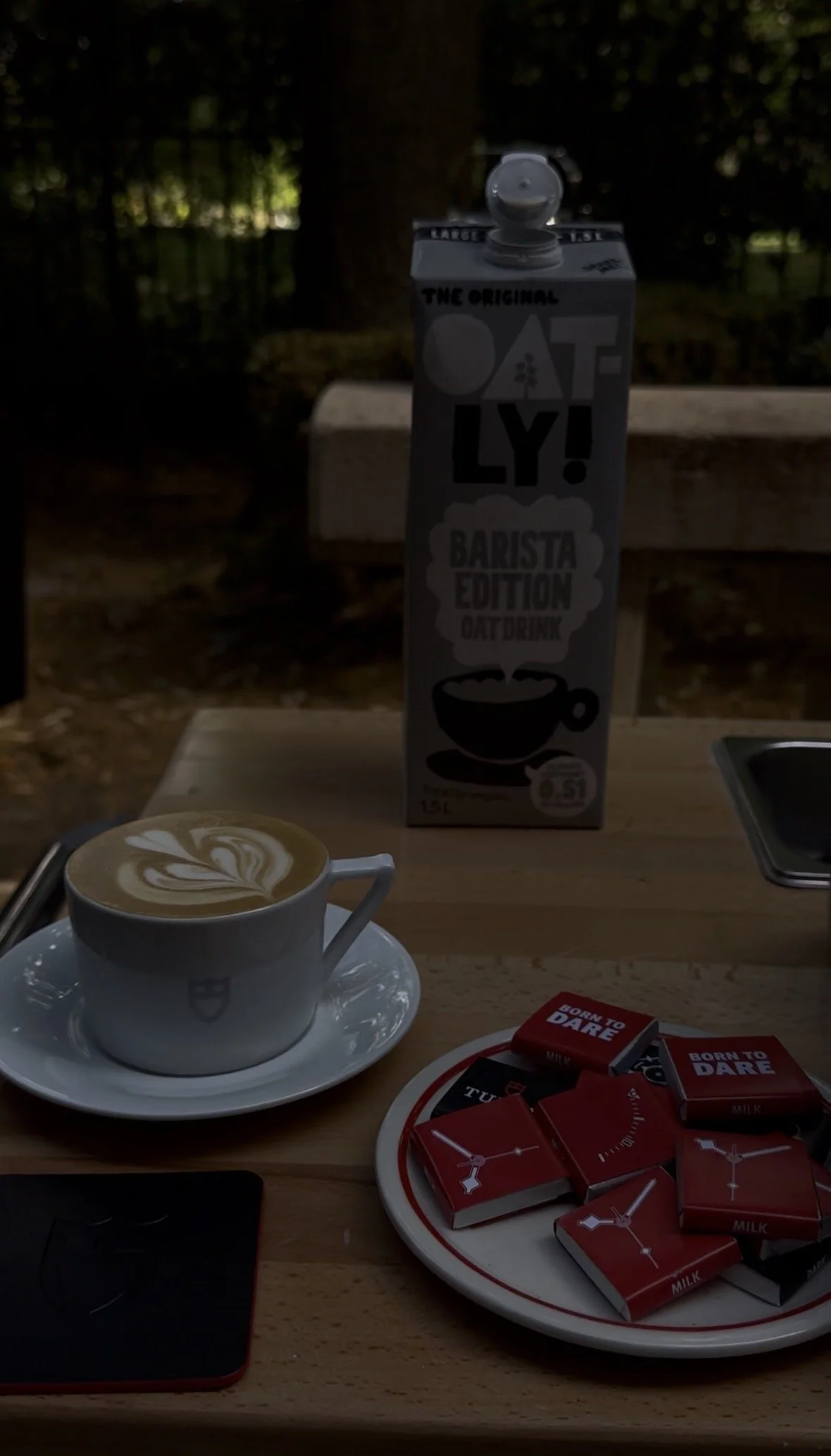 A cup of coffee with latte art on a saucer, a barista edition oat drink carton, a tray of chocolate bars labeled 'Born to Dare', and a smartphone on a wooden table outdoors.