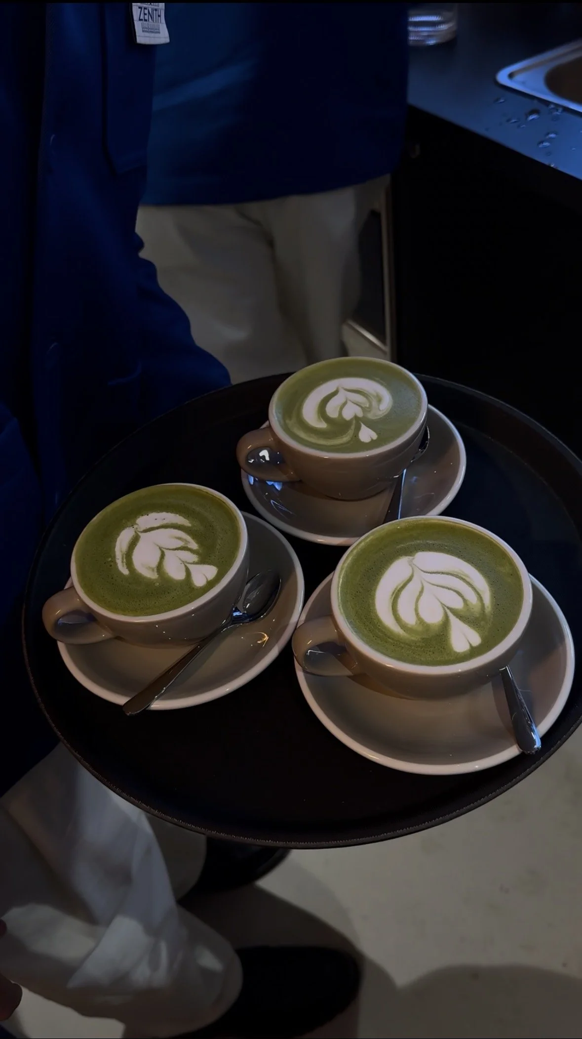Three cups of matcha latte with latte art on a round black tray, each cup on a saucer with a spoon, in a cafe or kitchen setting.