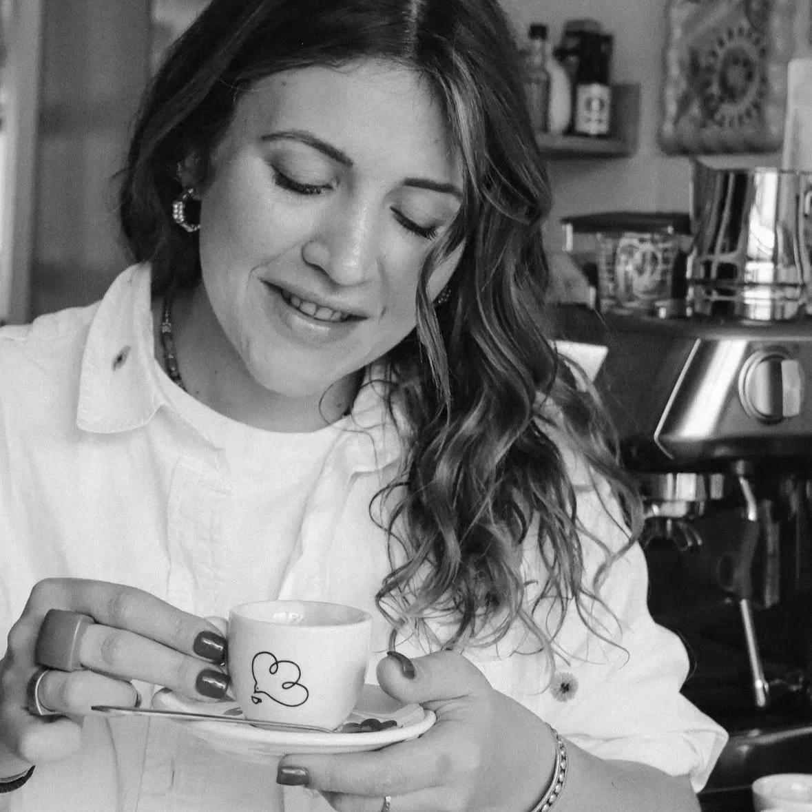 A woman with long wavy hair, earrings, and a white button-down shirt is holding a cup and saucer in a cozy kitchen.