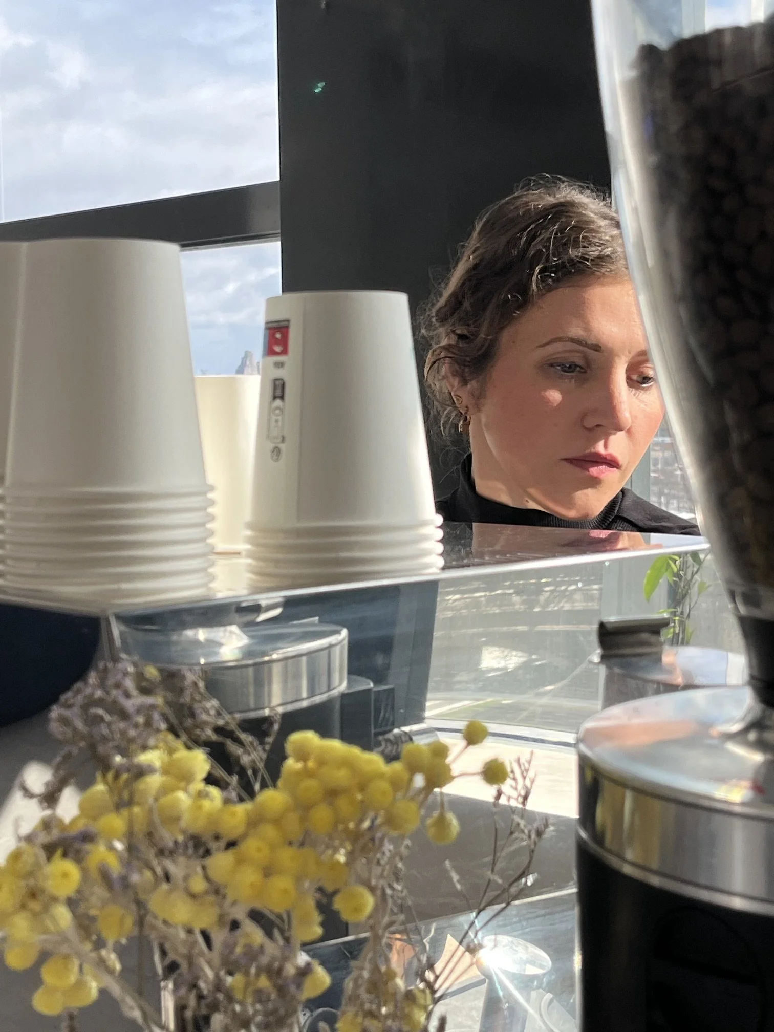 A woman with dark hair is looking down in front of a window with a view of a city building and cloudy sky. In the foreground, there are white disposable cups, a flower arrangement with yellow and white flowers, and a metal container.