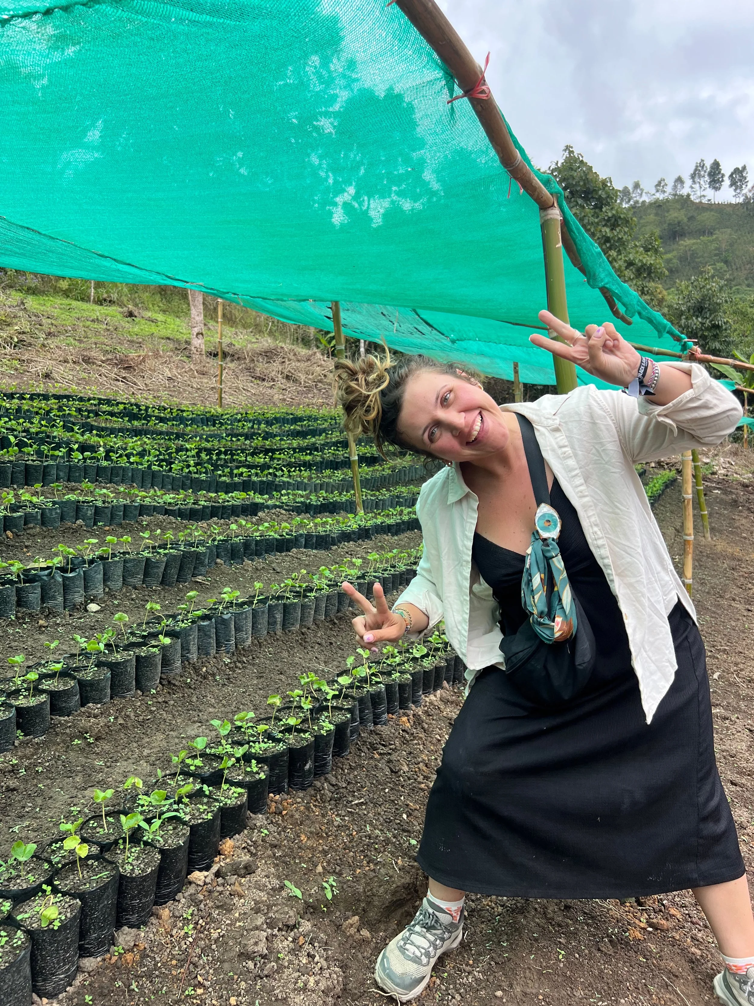 A woman making a peace sign while crouching in a farm or garden with rows of small green seedlings in black pots, covered by a green mesh shade cloth.