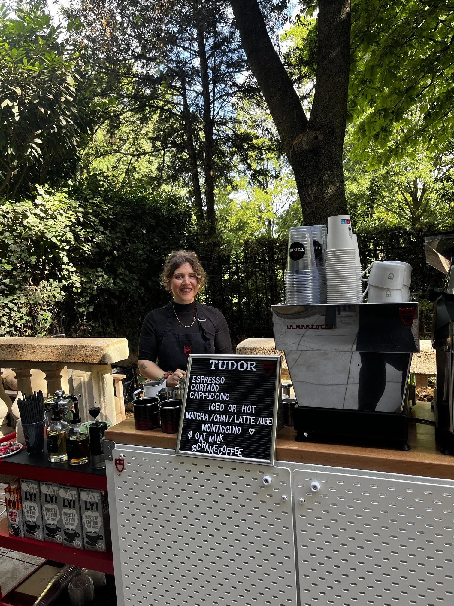 A woman standing behind a coffee stand outdoors surrounded by trees and green foliage. The stand displays several coffee cups, a sign listing coffee options, and a espresso machine.