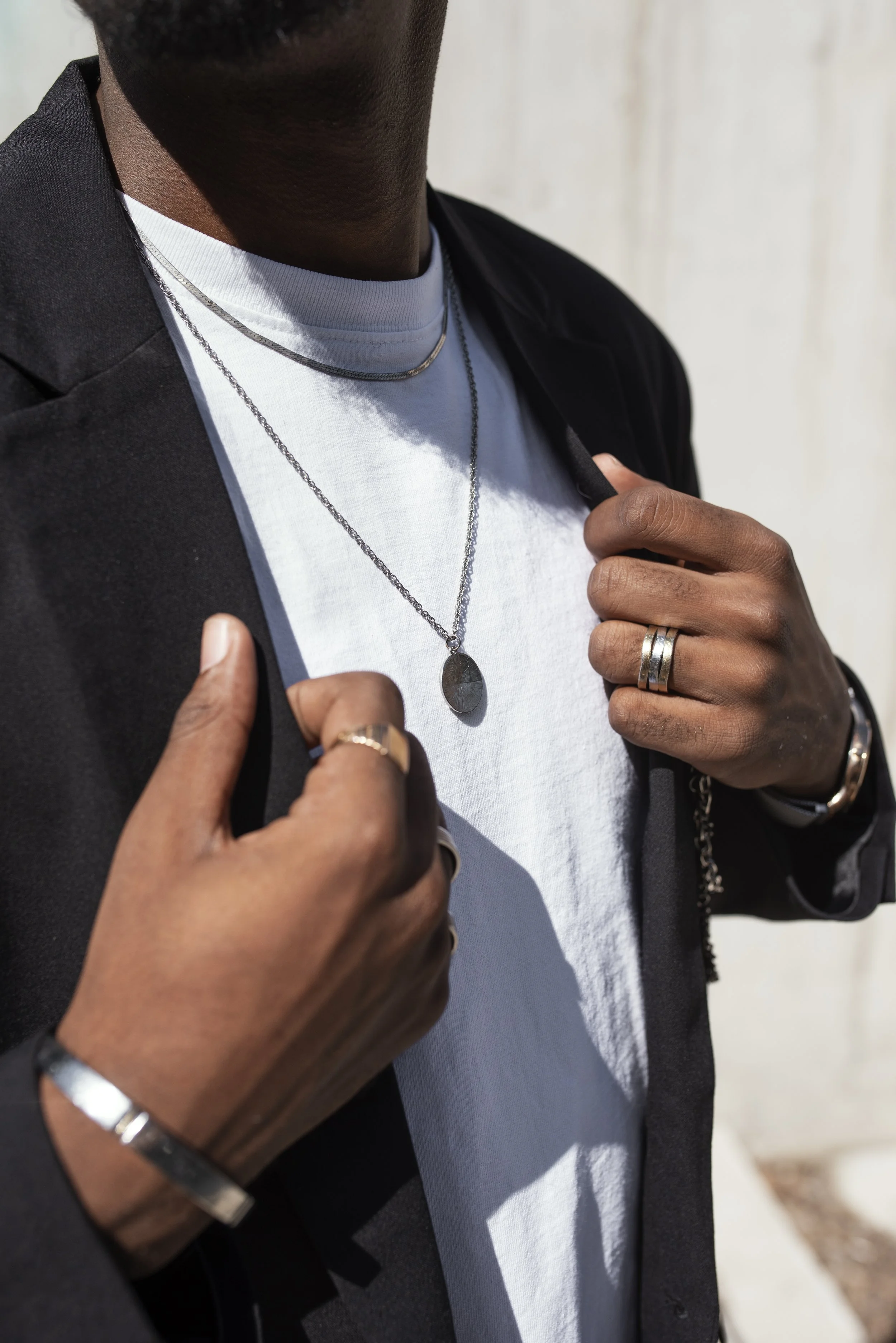 Close-up of a man wearing a black blazer, white t-shirt, layered silver necklaces, and rings on his fingers, holding his blazer lapel.