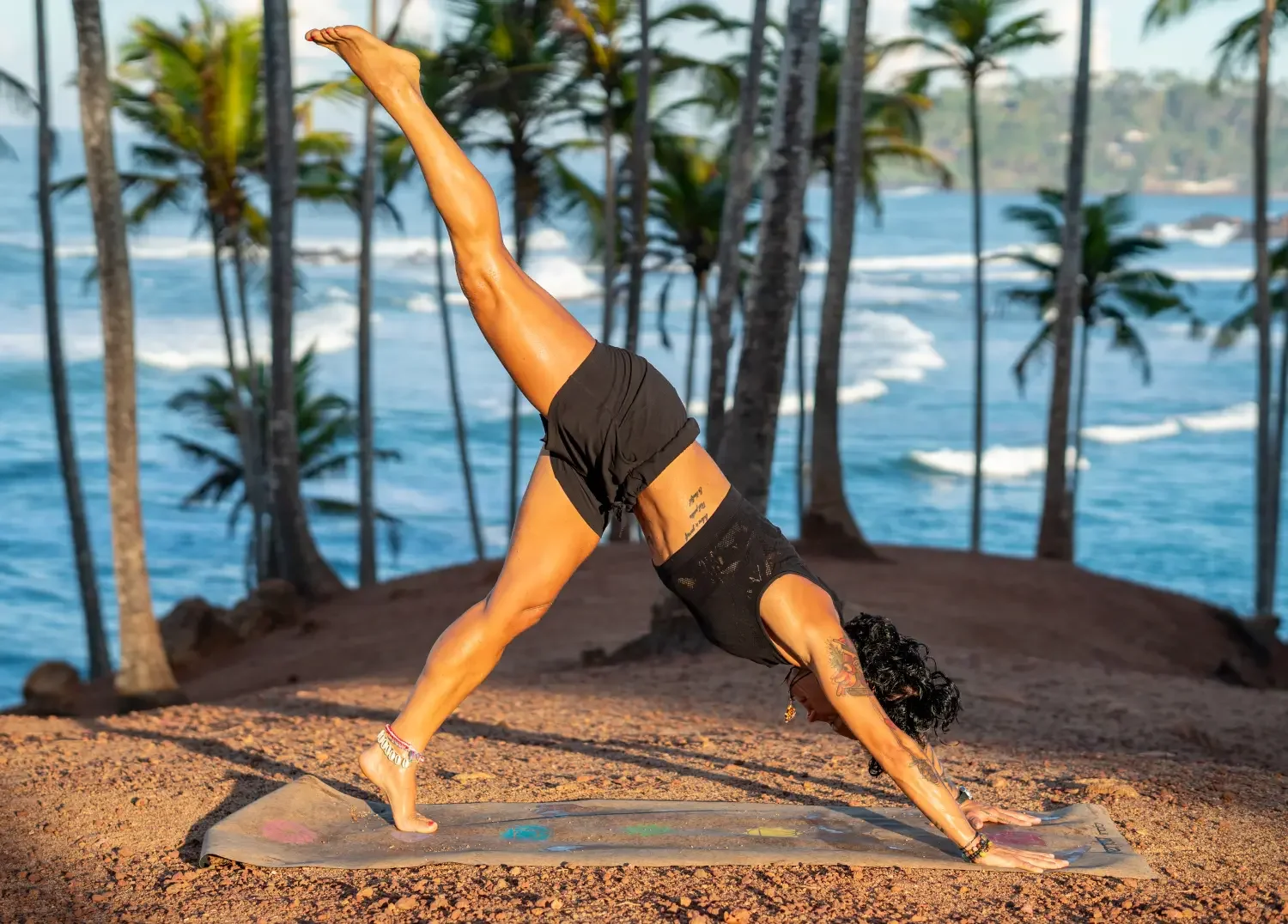 Caro in three-legged downward dog yoga pose on beach with palm tree backdrop