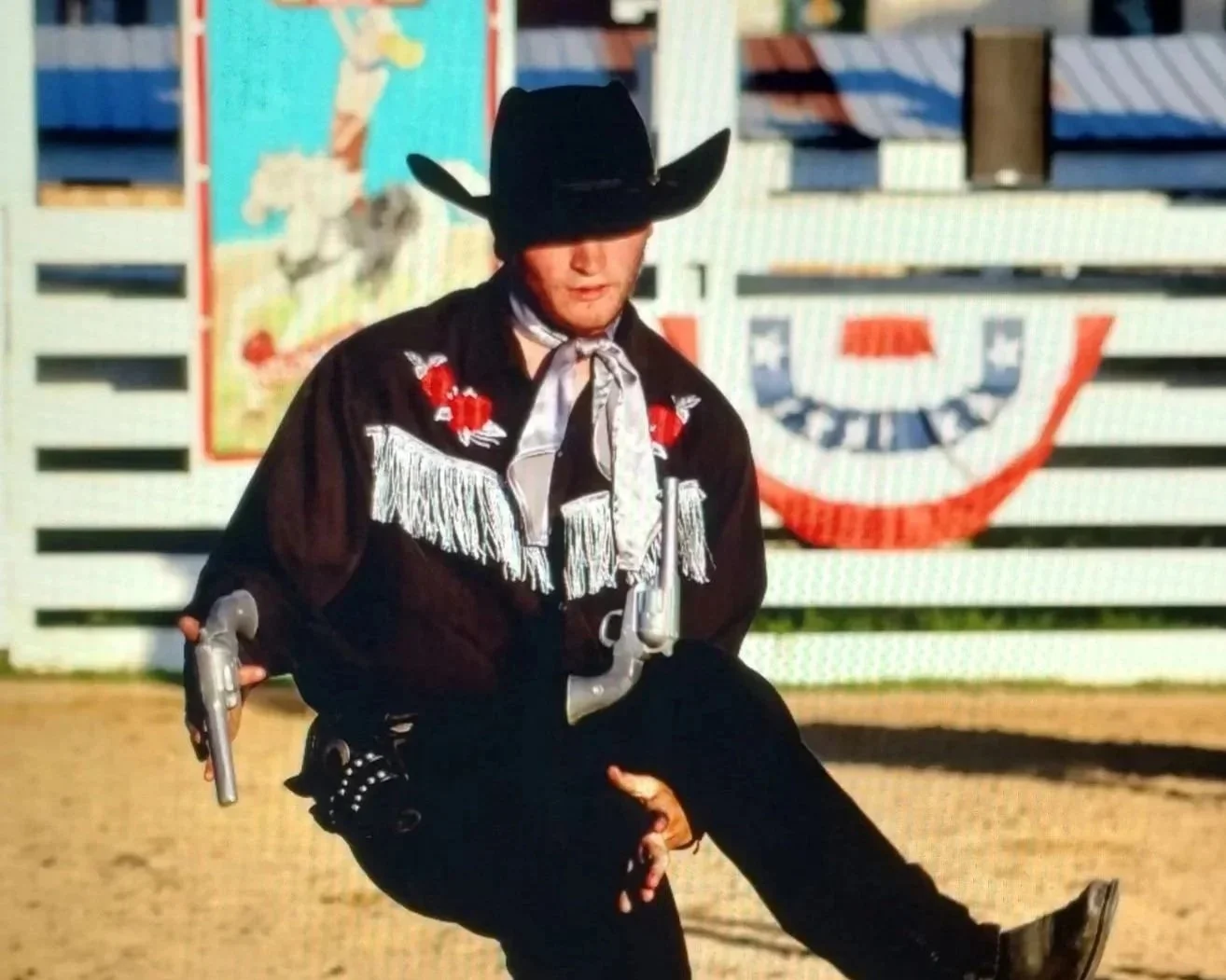 A person dressed as a sheriff or cowboy, wearing a large black hat, black fringe jacket, and bandana, holding two toy guns, in a rodeo or Western-themed setting.