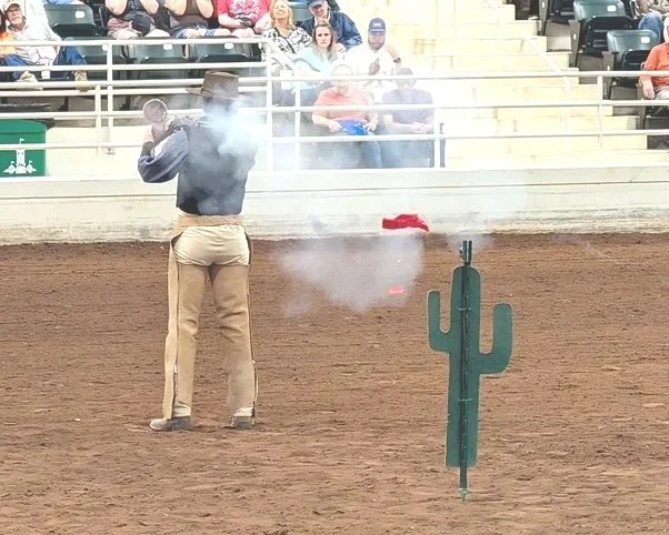 A person dressed in Western-style clothing stands in a dirt arena during a rodeo, holding a gun with smoke coming out of it, with a crowd watching from bleachers in the background, and a green artificial cactus in the foreground.