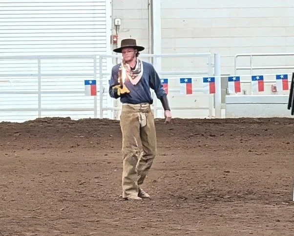A woman dressed in Western attire, including a wide-brimmed hat, bandana, and cowboy boots, standing on a dirt arena with Texas flags hanging in the background.
